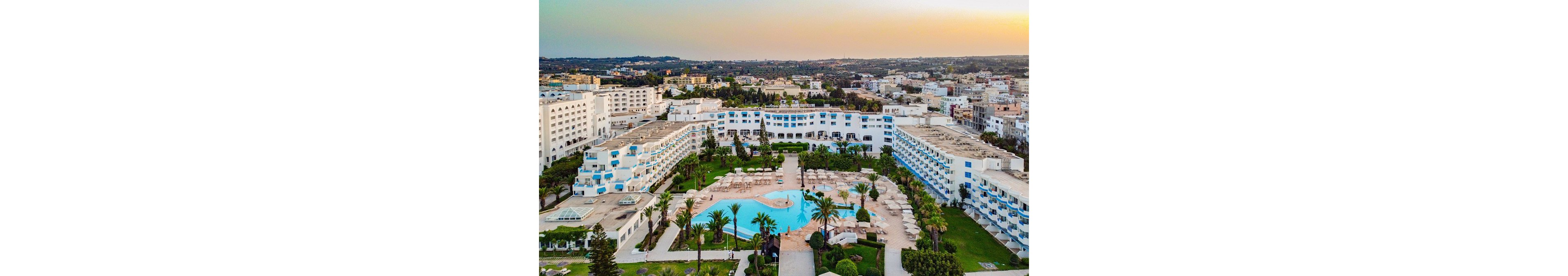 A large white resort hotel complex surrounds a central outdoor swimming pool. Numerous sun loungers and umbrellas are arranged neatly around the pool within landscaped green gardens and palm trees. The scene is captured from an elevated aerial perspective, showing the extensive hotel wings and surrounding urban area. Warm evening light creates a relaxed vacation atmosphere over the city and resort.