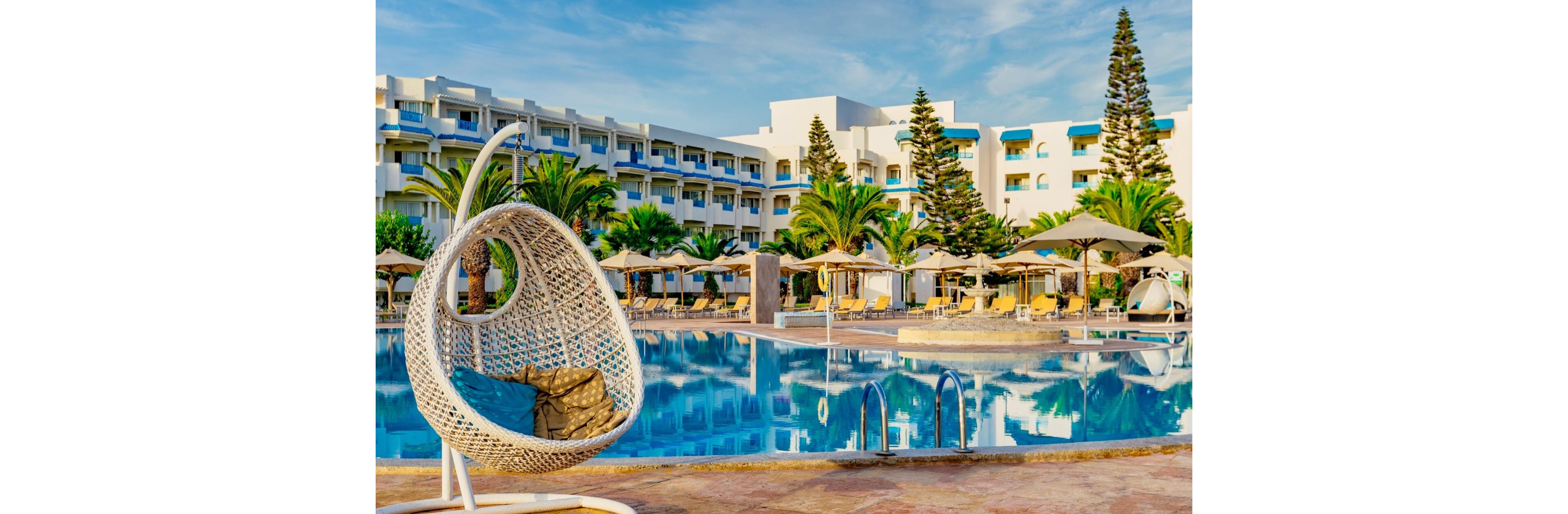 A large resort swimming pool reflects a multi-story hotel building in the background. A white woven hanging chair with cushions is positioned prominently at the poolside. Sun loungers and umbrellas line the far side of the pool, surrounded by palm trees and tall evergreens. The scene is brightly lit with clear blue skies, creating a relaxed vacation atmosphere.