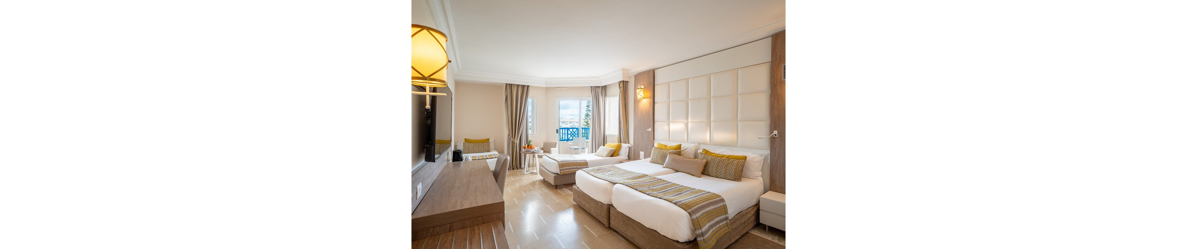 A spacious hotel room features a large double bed and an additional single bed with coordinated yellow and white bedding. The room includes a work desk, wall-mounted television, and contemporary lighting fixtures. Neutral beige tones dominate the decor, complemented by patterned cushions and throws. Large windows and a balcony door open to a view of the sea and blue balcony railings, creating an airy, relaxing atmosphere.