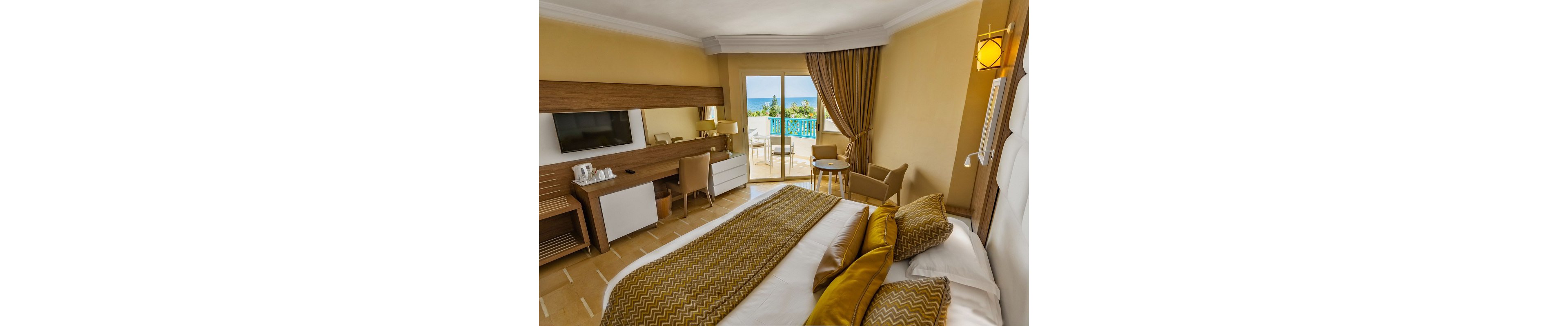 A modern hotel bedroom features a large neatly made bed with patterned yellow and white bedding. The room includes a wooden desk with a flat-screen TV, chairs, and a small table near the balcony doors. Warm yellow walls and curtains create a cozy, inviting atmosphere. Through the glass doors, a balcony with blue railing overlooks the sea and clear sky.