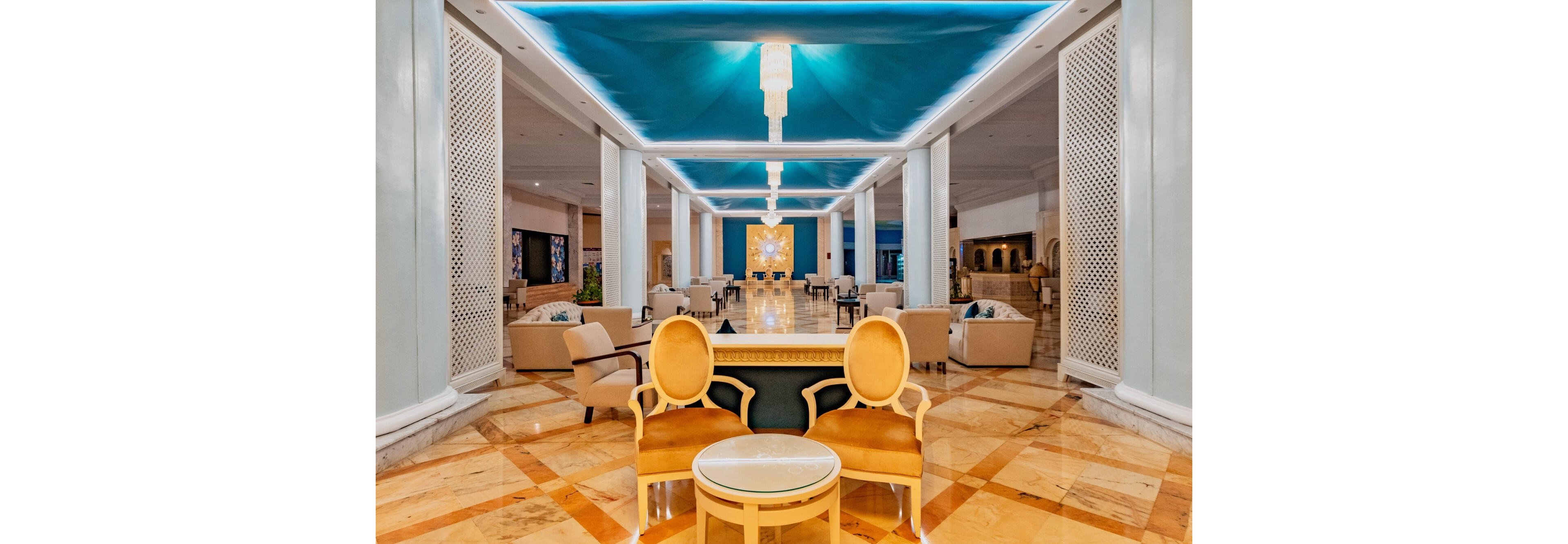 Spacious hotel lobby interior featuring two ornate yellow chairs and a small round table in the foreground. The space extends into a long corridor of seating areas with armchairs and sofas arranged symmetrically. A striking blue ceiling with recessed lighting and chandeliers contrasts with the warm marble floor. The overall atmosphere is upscale, bright, and welcoming, suggesting a luxury resort or grand hotel reception area.