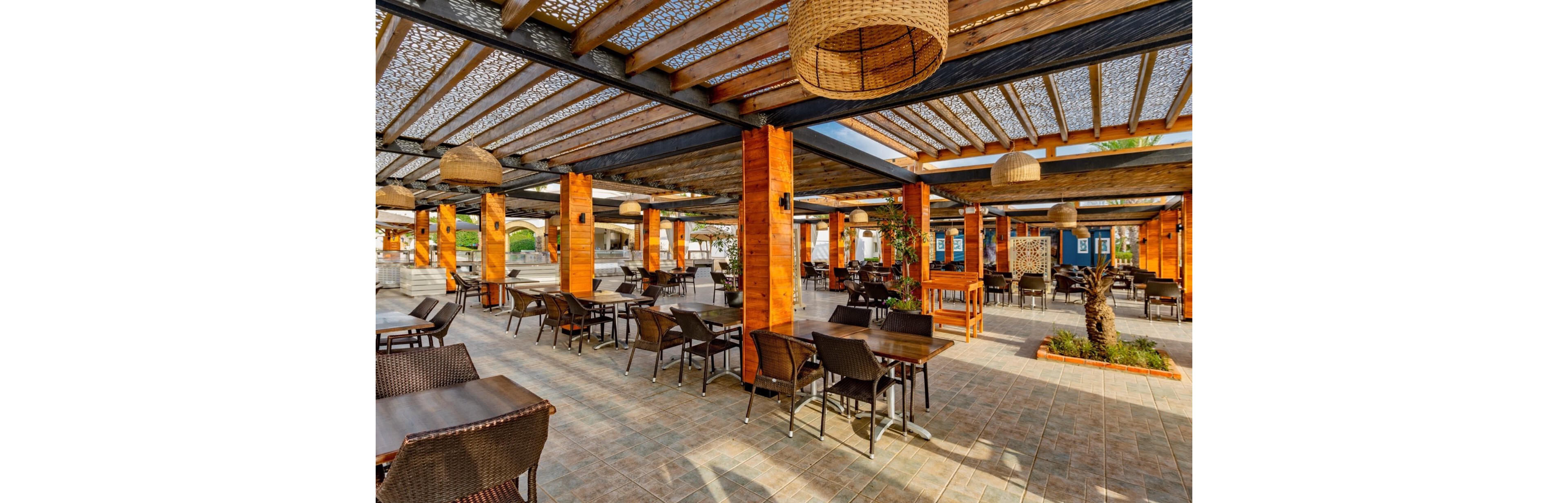 An open-air café terrace features rows of wooden pergola columns and wicker chairs arranged around dark tables. The setting appears to be part of a resort or hotel, with a tiled floor and shaded seating areas. Decorative woven pendant lights hang from the ceiling structure, casting patterned shadows. The space is empty of people, emphasizing the relaxed, sunny atmosphere and inviting outdoor dining experience.