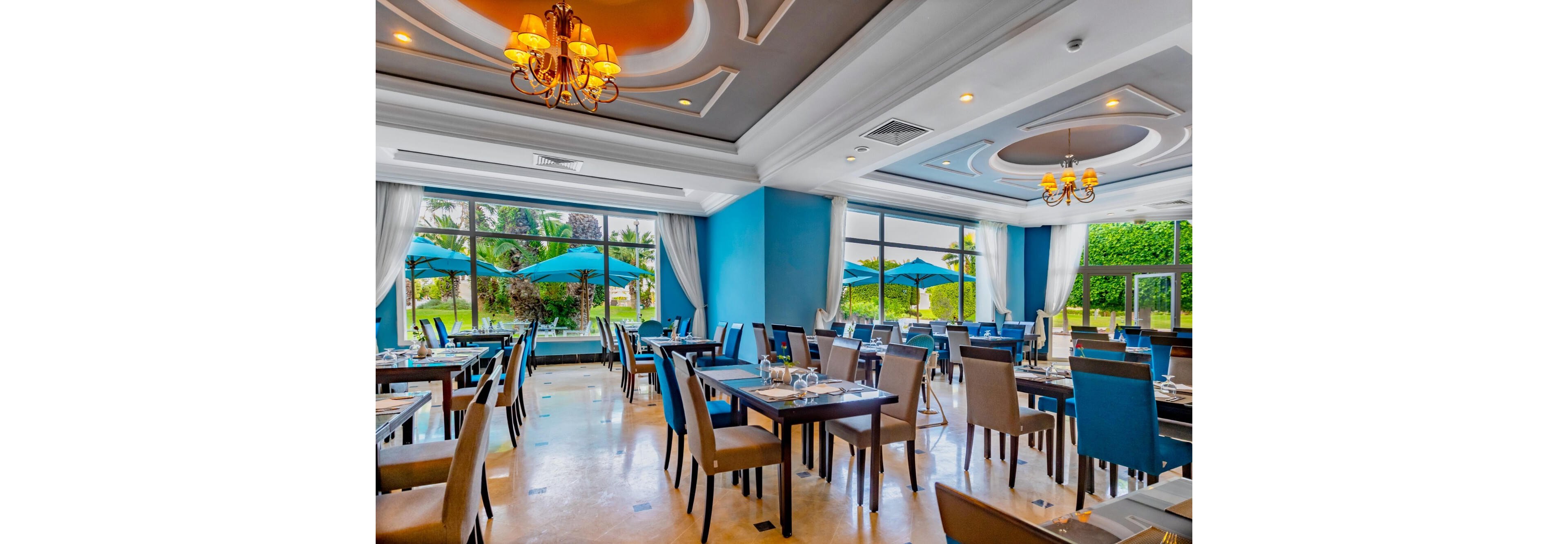 A spacious, contemporary restaurant dining room features neatly arranged tables and chairs. Large floor-to-ceiling windows reveal a lush garden with palm trees and blue patio umbrellas outside. The interior decor includes blue accent walls, elegant chandeliers, and white coffered ceilings. Place settings are prepared on the tables, suggesting readiness for guests in a calm, inviting atmosphere.