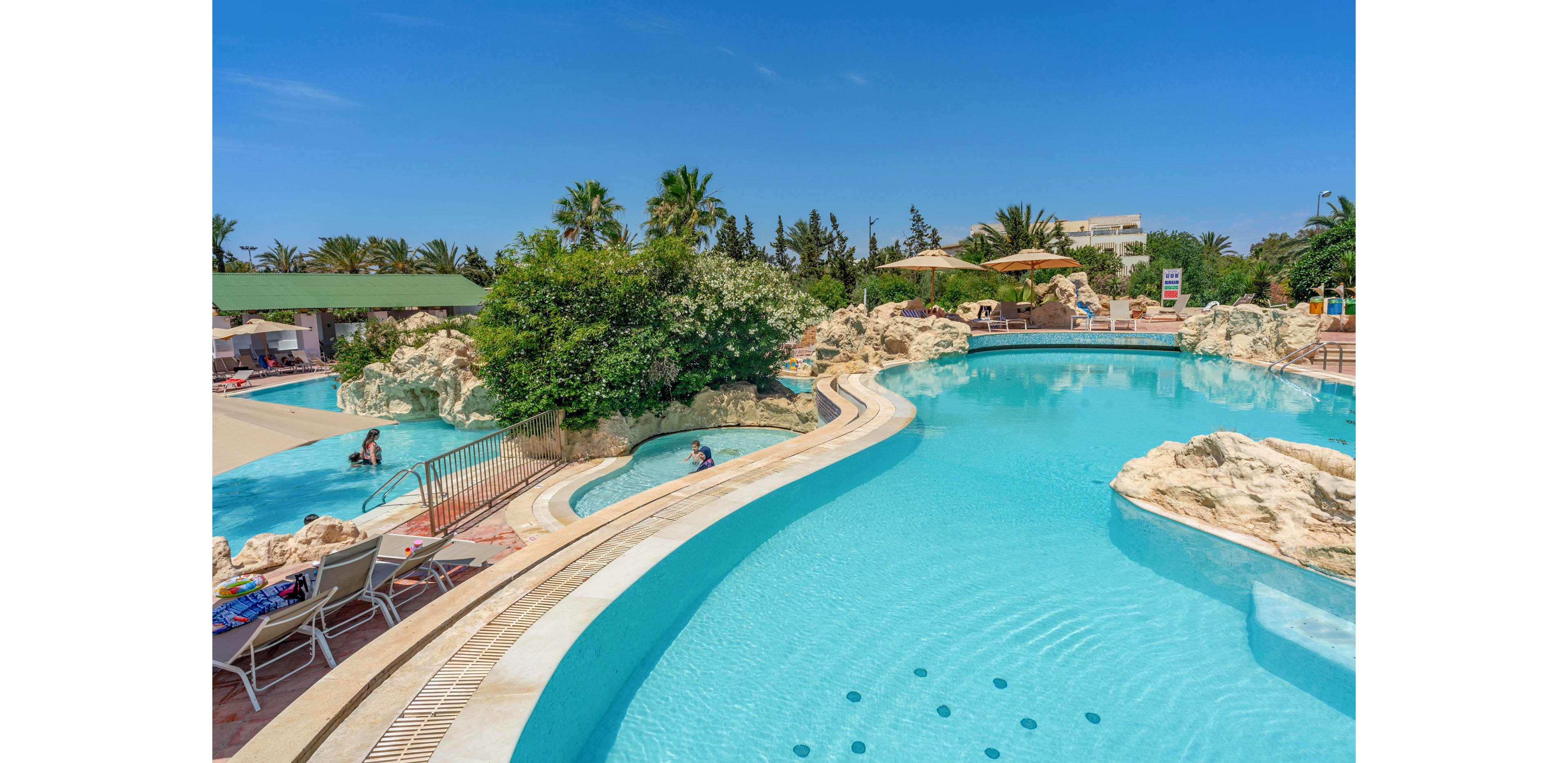 A large resort-style swimming pool curves around rocky formations and lush greenery under a clear blue sky. Guests relax in the water and on lounge chairs beside the pool. Palm trees and landscaped gardens surround the area, creating a tropical vacation atmosphere. Sun umbrellas and seating areas provide shaded spots for relaxation near the water.