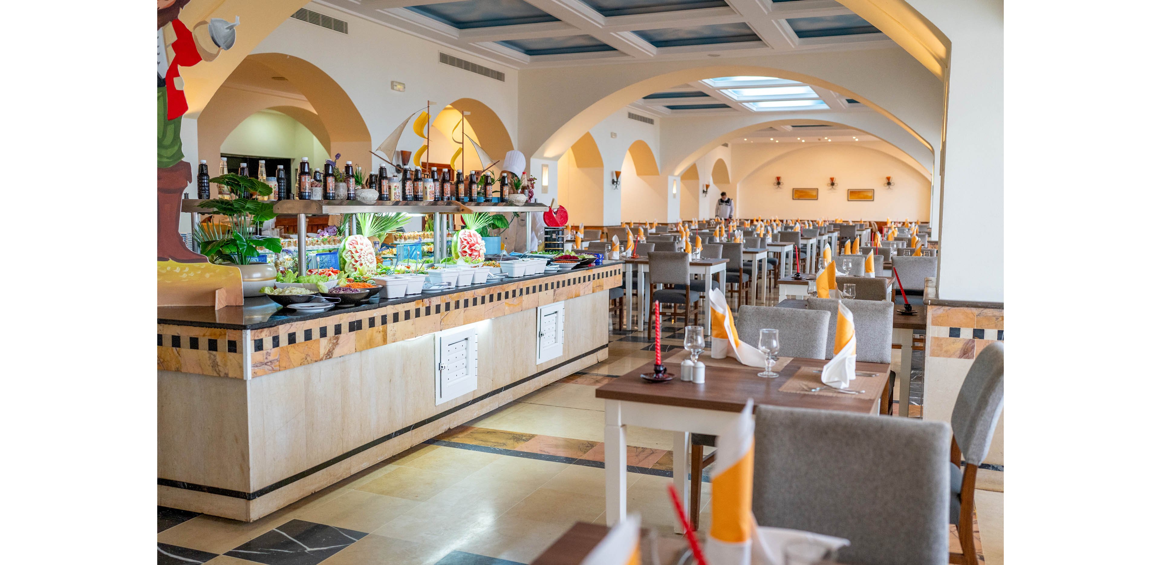 A spacious hotel restaurant features a central buffet counter filled with fresh salads and colorful dishes. Neatly arranged dining tables with folded yellow and white napkins and glassware line the room. Arched ceilings, warm lighting, and tiled floors create an inviting Mediterranean-inspired atmosphere. The restaurant appears ready for guests, with no people present and a clean, orderly setting.