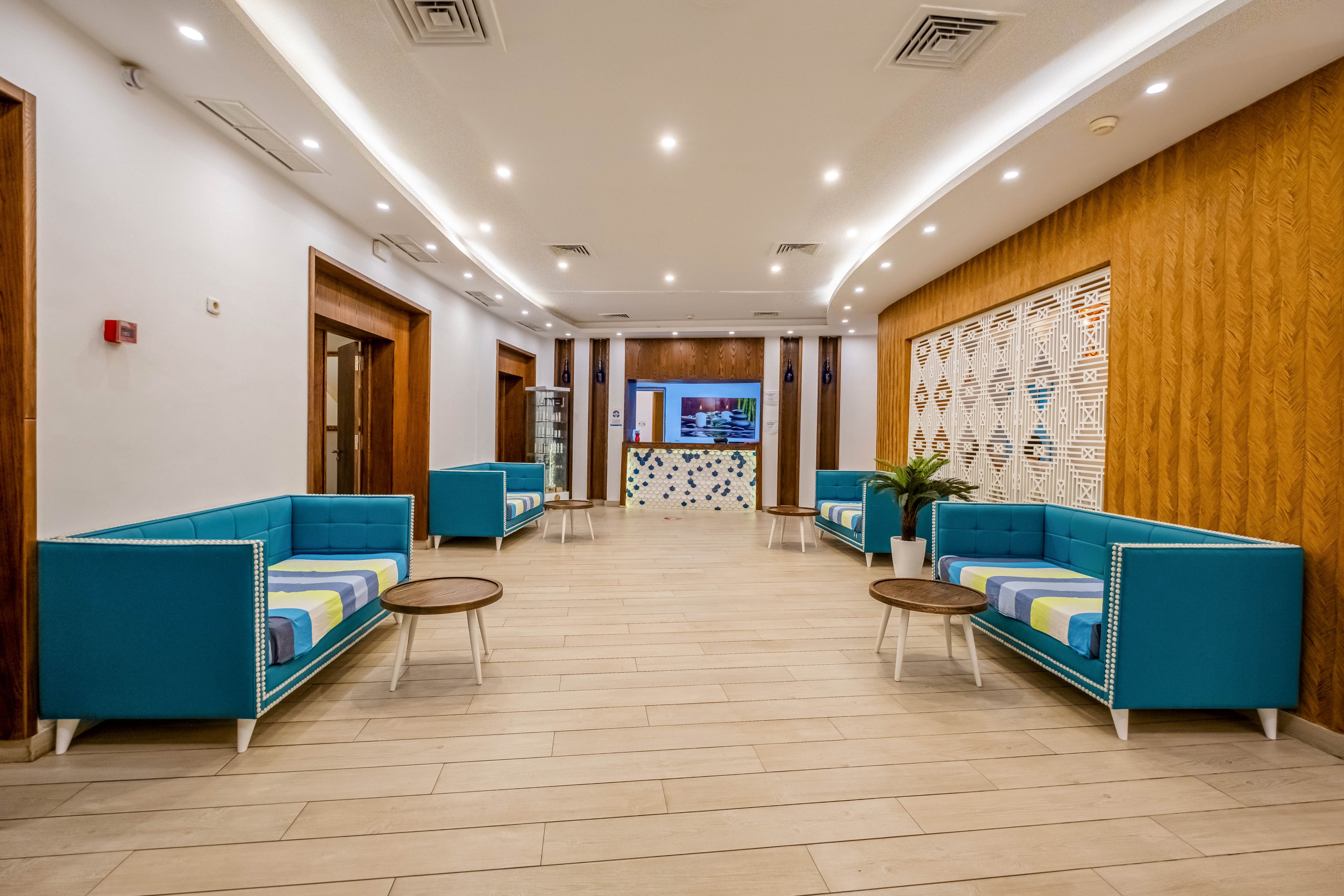 A contemporary lobby features teal sofas arranged symmetrically along a wide corridor. Light wood flooring and wooden wall panels create a warm contrast to the bright blue upholstery. Recessed ceiling lights and cove lighting illuminate the space evenly, leading the eye toward a reception desk and wall-mounted screen at the far end. Decorative patterned panels and indoor plants add visual interest and a welcoming atmosphere.