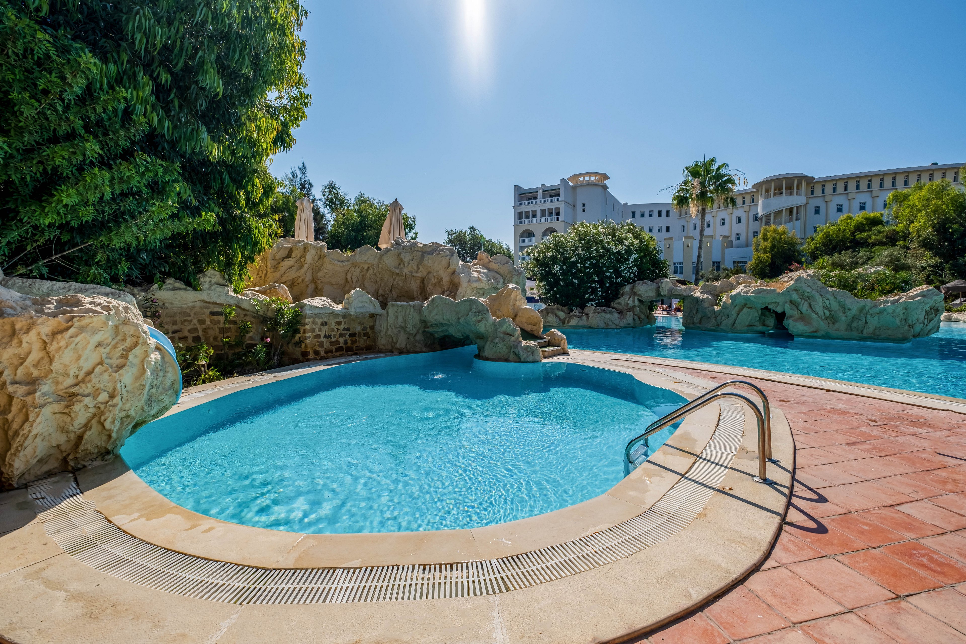 A bright outdoor swimming pool is set within a resort featuring natural rock formations and lush greenery. Clear blue water fills the curved pool, with a metal handrail leading down the steps. In the background, a larger pool area and a grand hotel building are visible under a clear blue sky. The scene suggests a relaxing vacation atmosphere in warm, sunny weather.