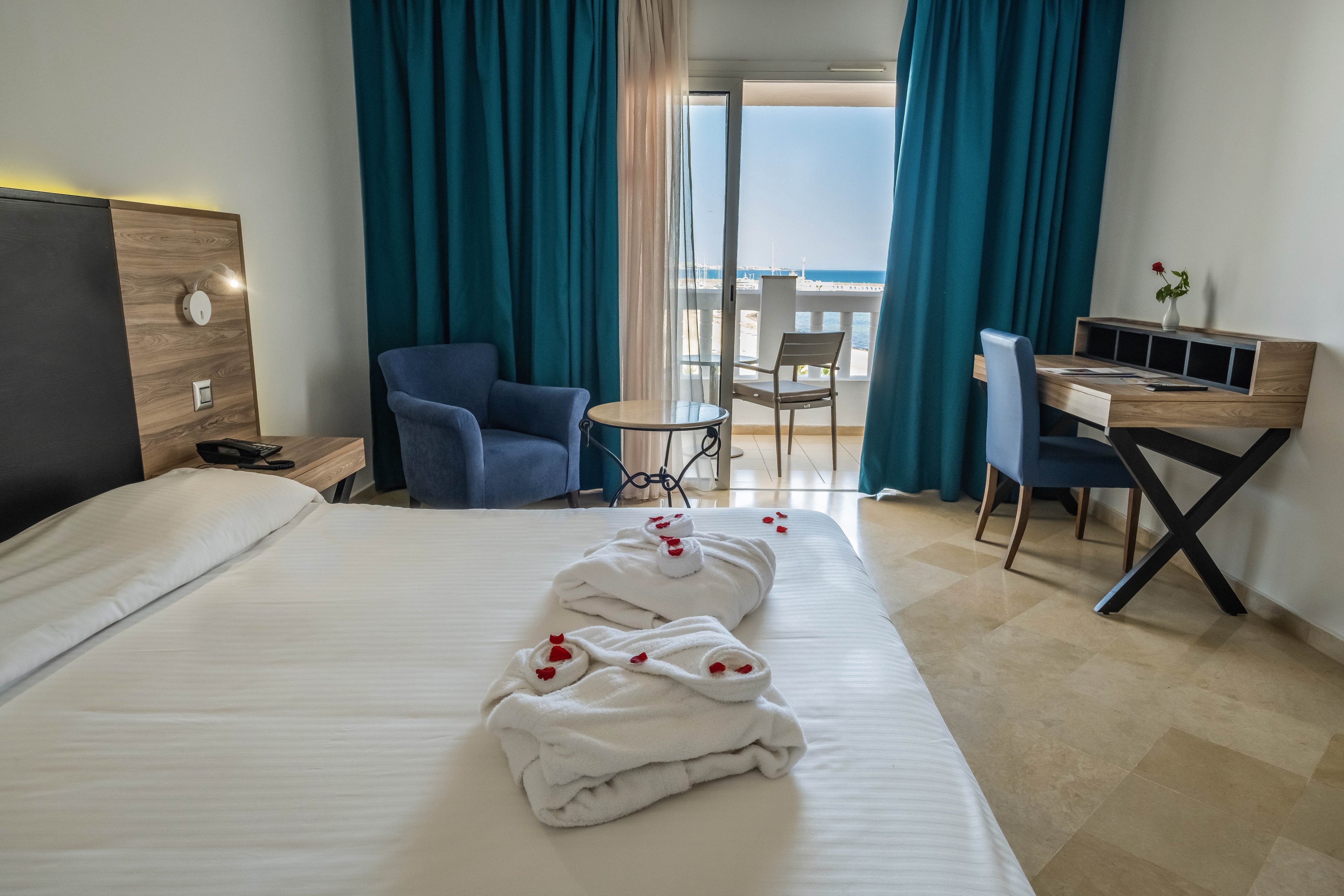 A neatly made bed with folded white robes and rose petals is the focal point of this modern hotel room. The space includes a wooden headboard, a small seating area with blue armchairs, and a compact writing desk. Floor-to-ceiling curtains frame a glass door that opens onto a balcony with chairs and a view of the sea. Soft natural light and neutral tones create a calm, inviting atmosphere suitable for vacation or business travel.