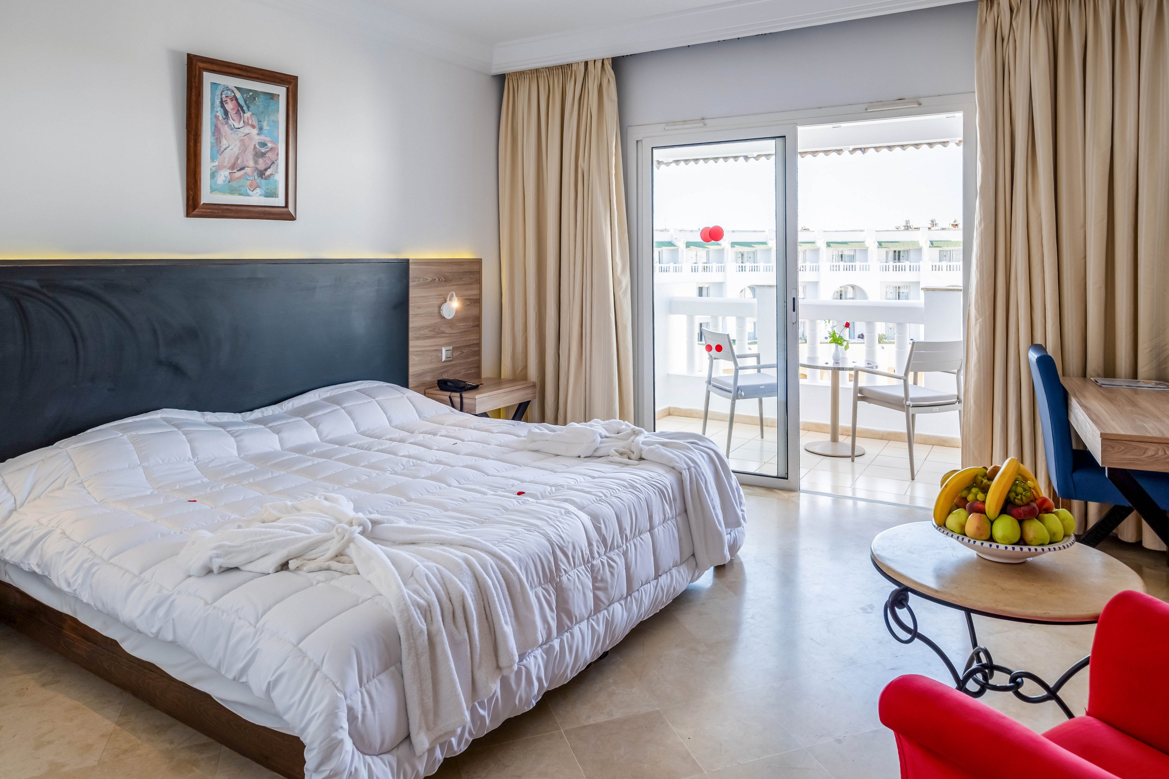 A neatly made double bed dominates a bright hotel bedroom with light walls and a dark headboard. The room opens onto a balcony through large glass doors, revealing outdoor chairs and a view of neighboring buildings. A small round table holds a bowl of fresh fruit, and a desk with a chair sits near the window. Warm curtains, a framed artwork above the bed, and soft lighting create a clean, inviting atmosphere.