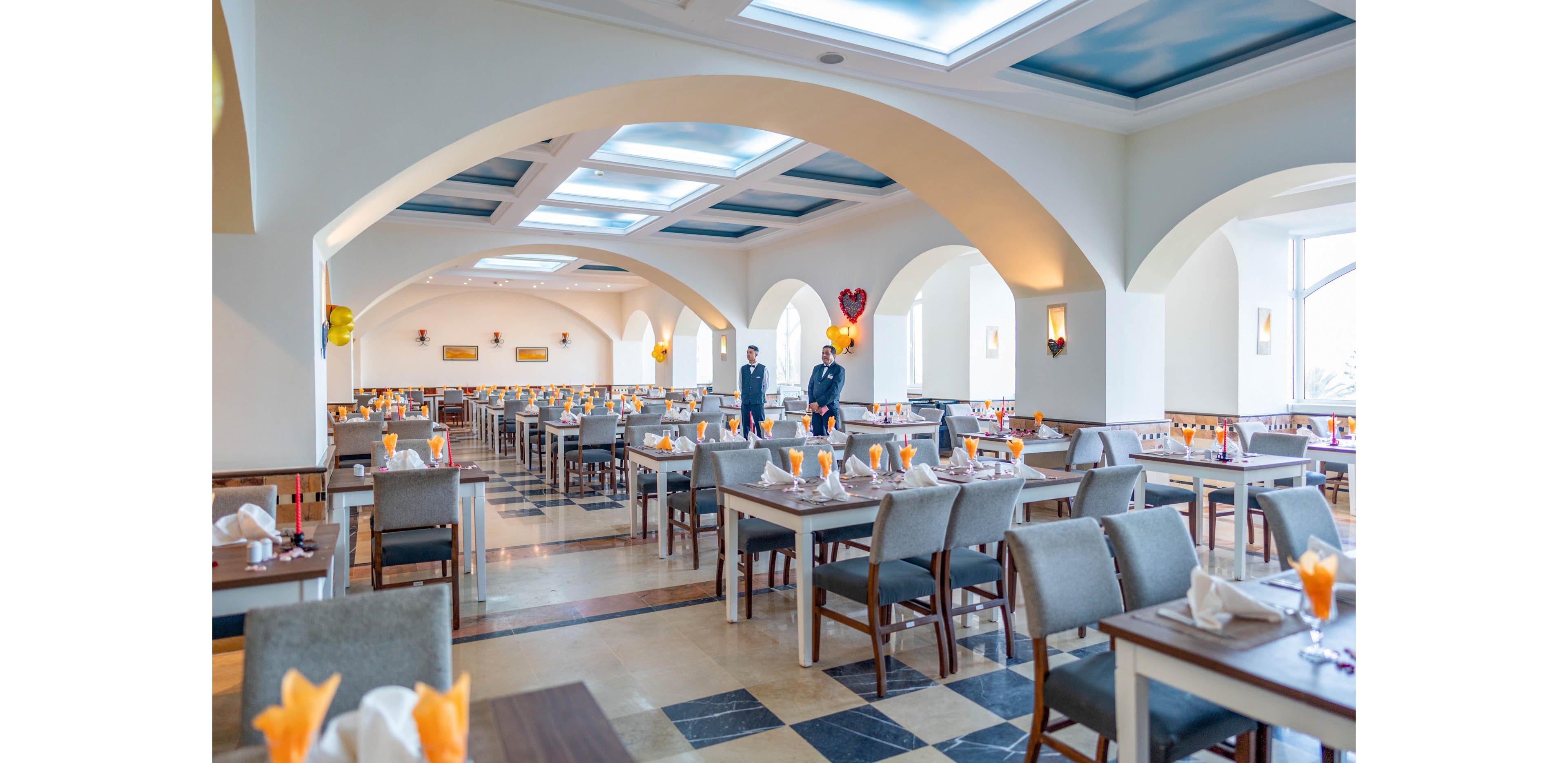 A spacious restaurant dining hall features neatly arranged tables and chairs under arched ceilings and skylights. Each table is set with folded napkins and orange-colored drinks, ready for guests. Two formally dressed staff members stand in the center of the room, adding a professional touch. Warm lighting and decorative balloons create a welcoming, celebratory atmosphere.