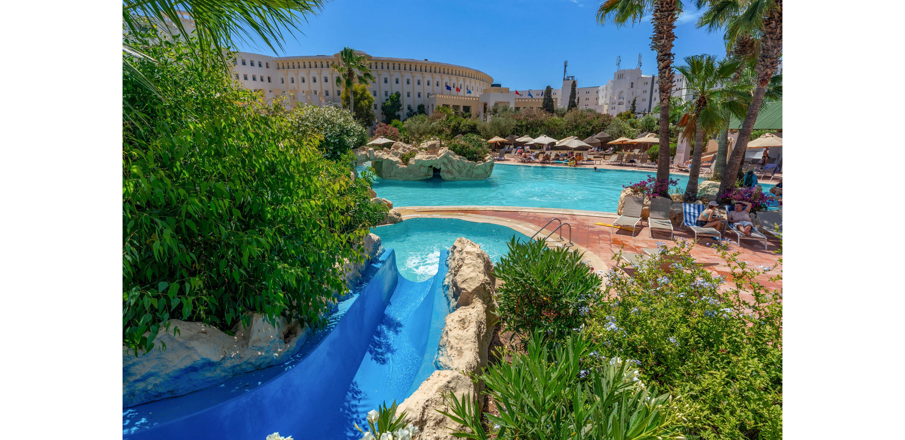 A large resort swimming pool is surrounded by lush greenery and tall palm trees. Guests relax on sun loungers under umbrellas while others stand in the shallow water. A bright blue waterslide curves through rocks into the pool, adding a playful element to the scene. In the background, a grand hotel building with multiple windows and flags rises above the pool area under a clear blue sky.