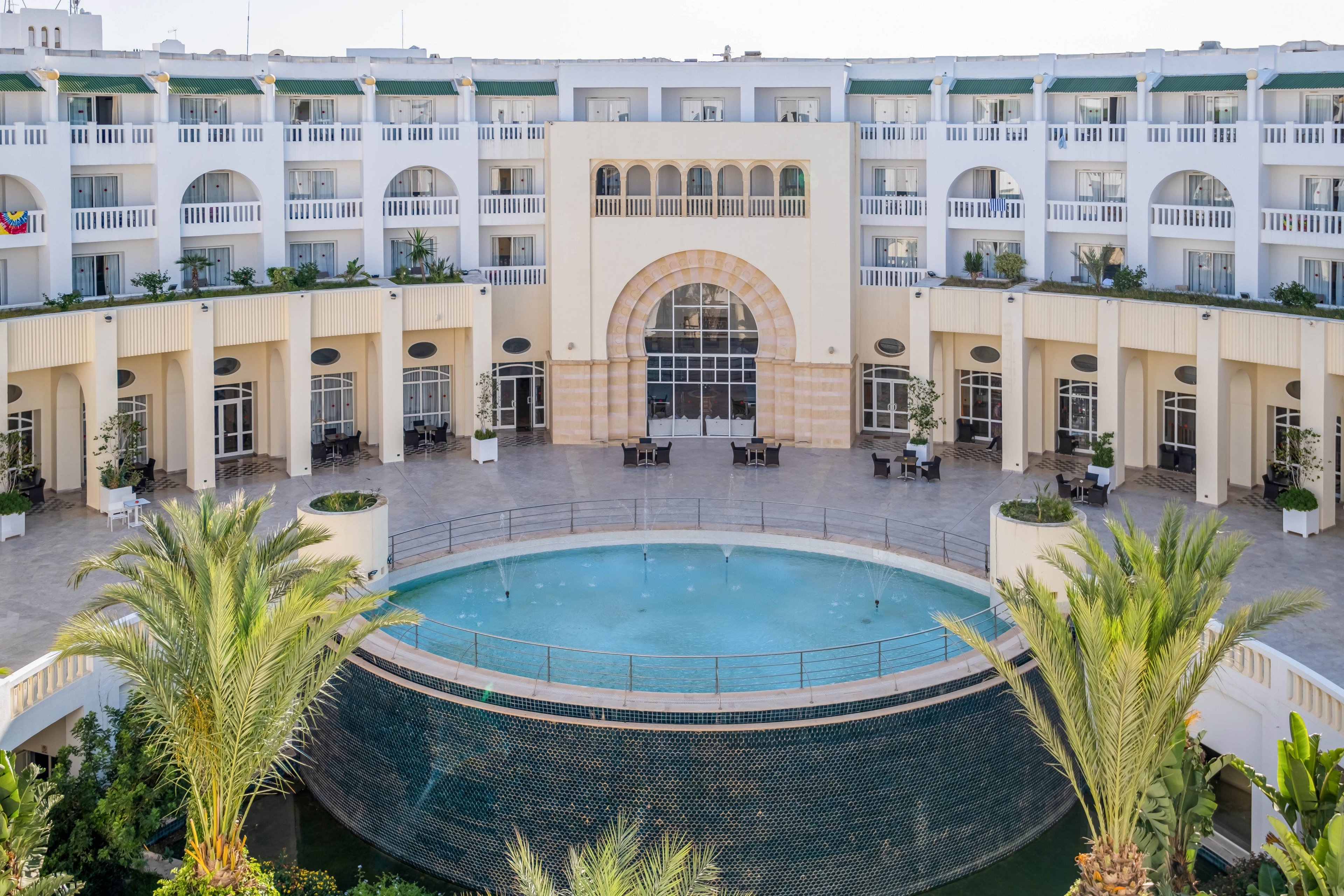 A large, elegant hotel building surrounds a circular courtyard with a central fountain pool. The architecture features multiple stories of white arches, balconies, and tall windows. Palm trees and greenery frame the foreground, adding a tropical touch to the scene. Outdoor seating and glass doors suggest a welcoming space for guests to relax and socialize.