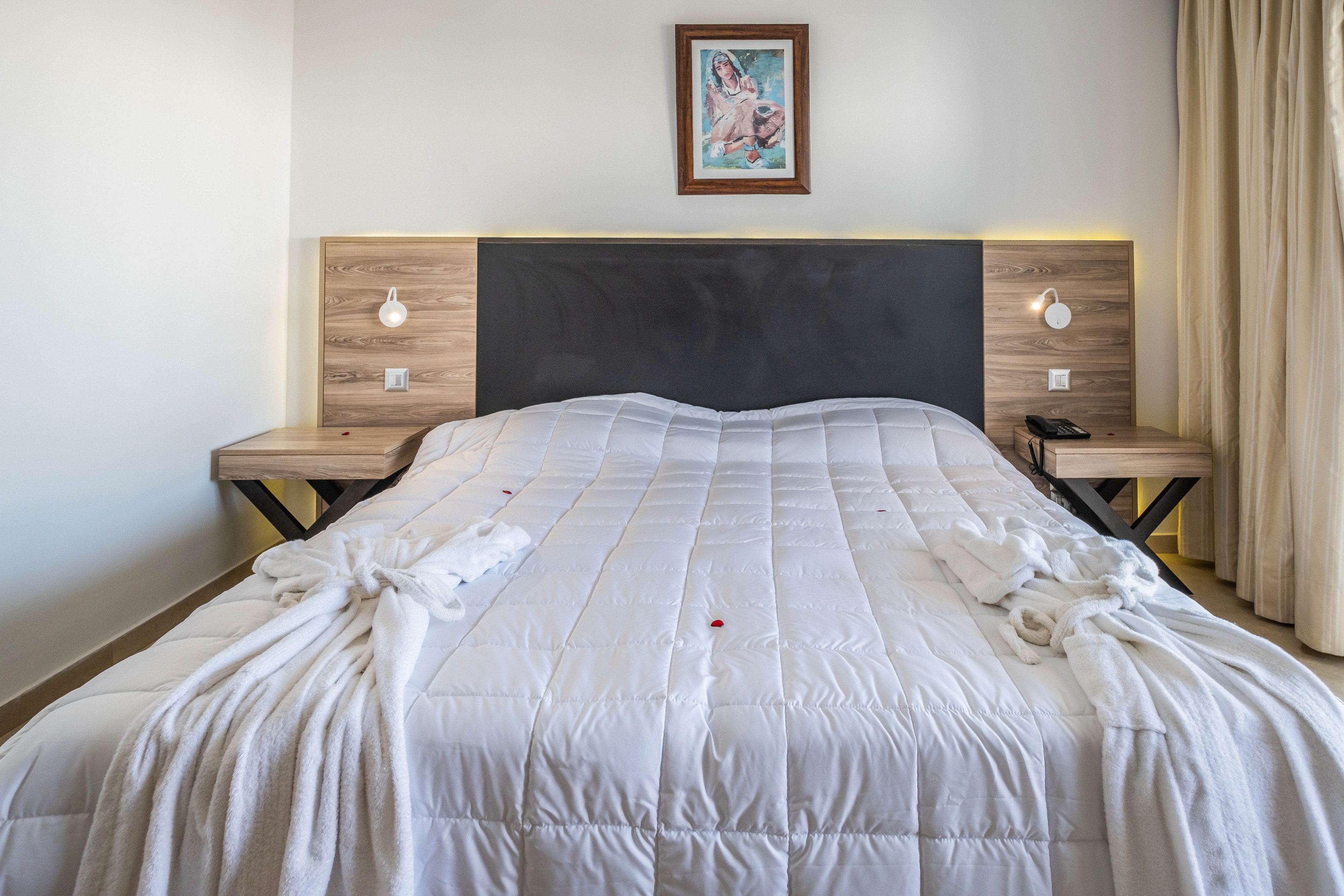 A neatly made double bed with a white quilted comforter is centered in a modern bedroom. Wooden bedside tables with built-in wall lamps flank the bed, and a black headboard contrasts with the light walls. A framed artwork hangs above the headboard, adding a decorative focal point. Soft natural light enters from a curtained window on the right, creating a calm and inviting atmosphere.
