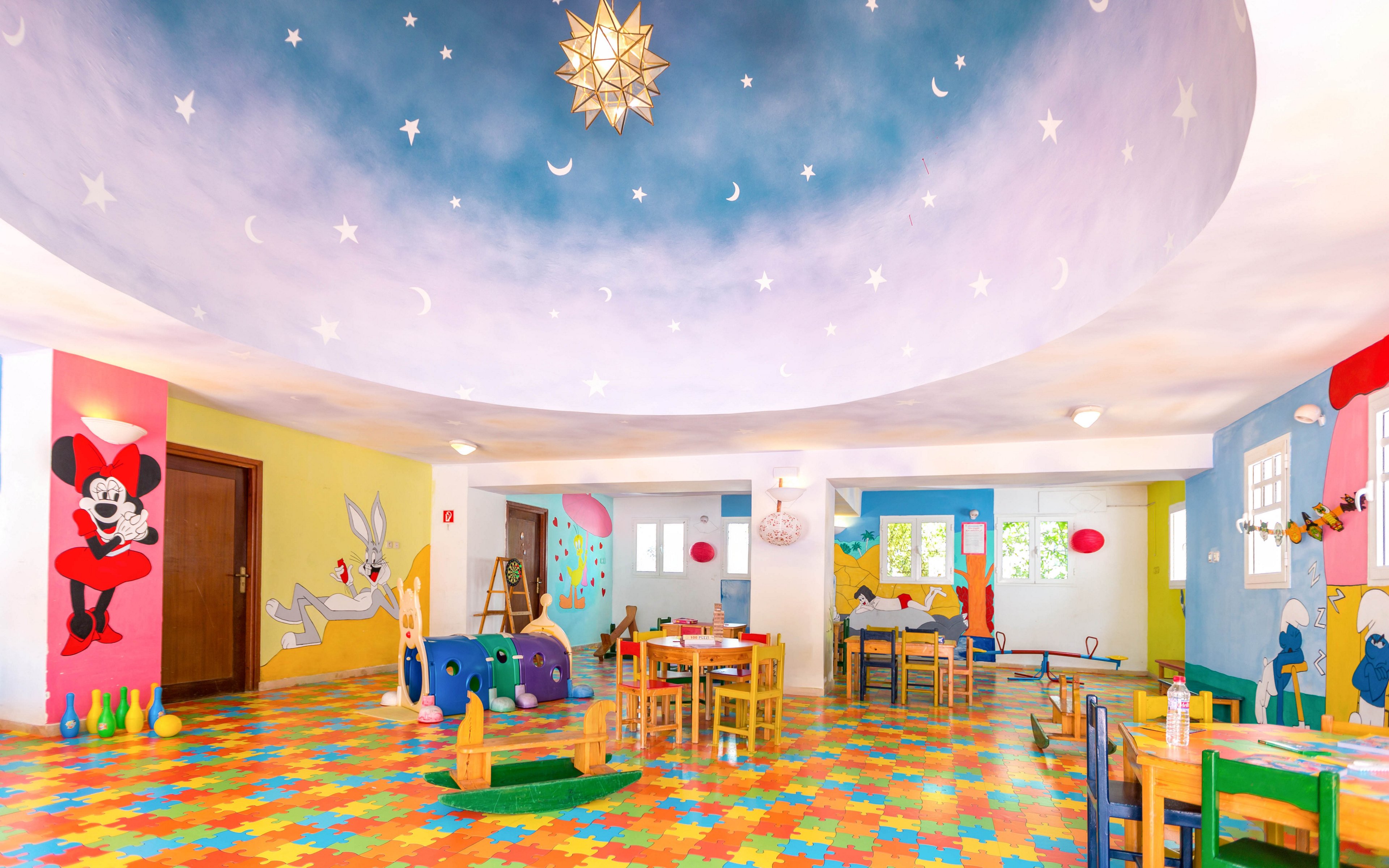 A bright, colorful kindergarten playroom features children and an adult engaging in activities at small tables. The room is decorated with cartoon characters on the walls and a painted ceiling showing stars, moons, and a gradient sky. The floor is covered with interlocking puzzle mats in vibrant colors, and various toys and play equipment are scattered around. Large windows and interior lights create a cheerful, welcoming atmosphere for play and learning.
