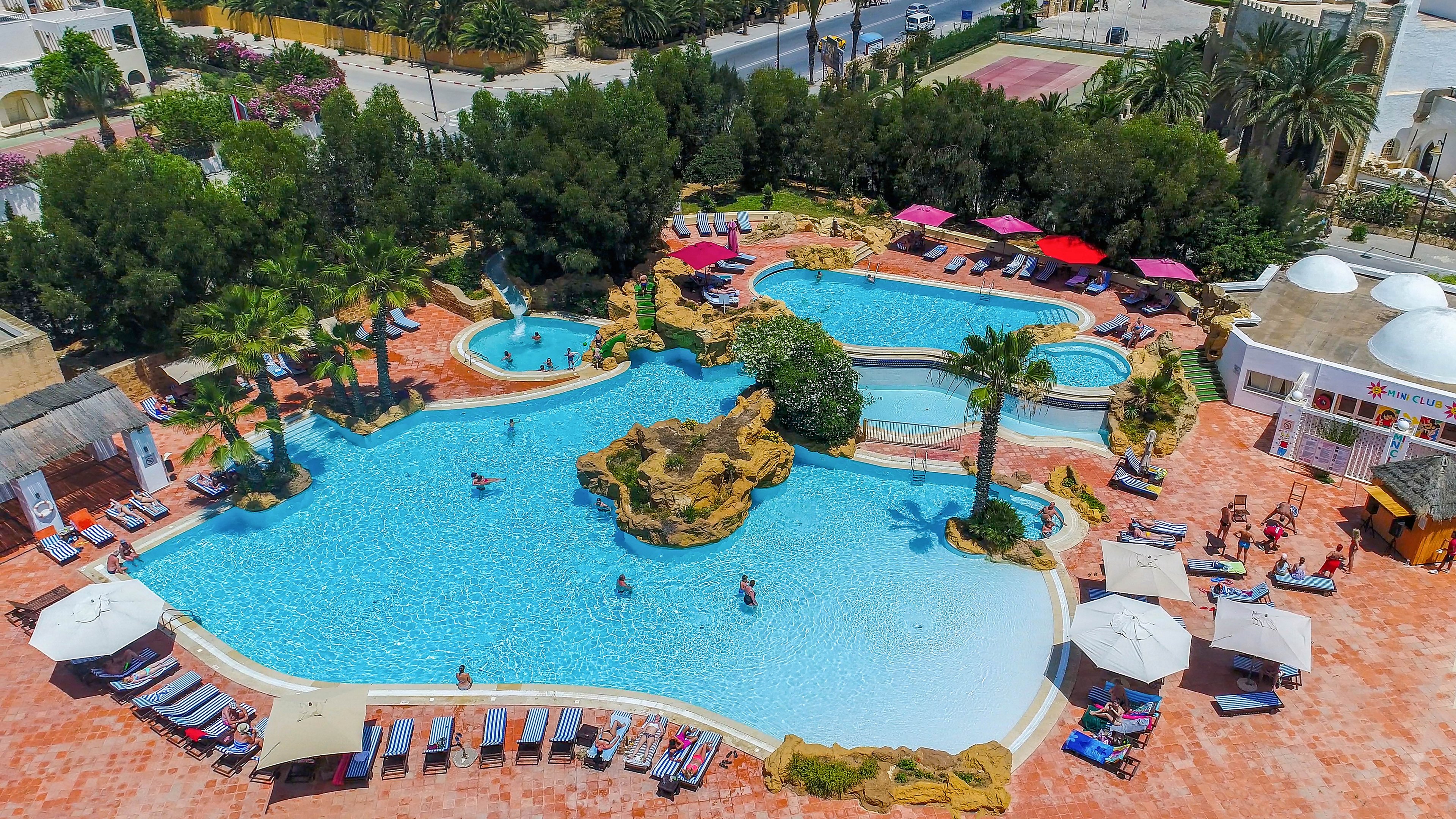 An aerial view showcases a large resort-style swimming pool complex with curved, lagoon-like shapes. Guests relax on sun loungers around the terracotta-tiled deck while others swim and play in the clear blue water. Rock formations, palm trees, and greenery create a tropical atmosphere, and several red umbrellas provide shaded seating areas. In the background, additional buildings and trees frame the tranquil vacation setting.