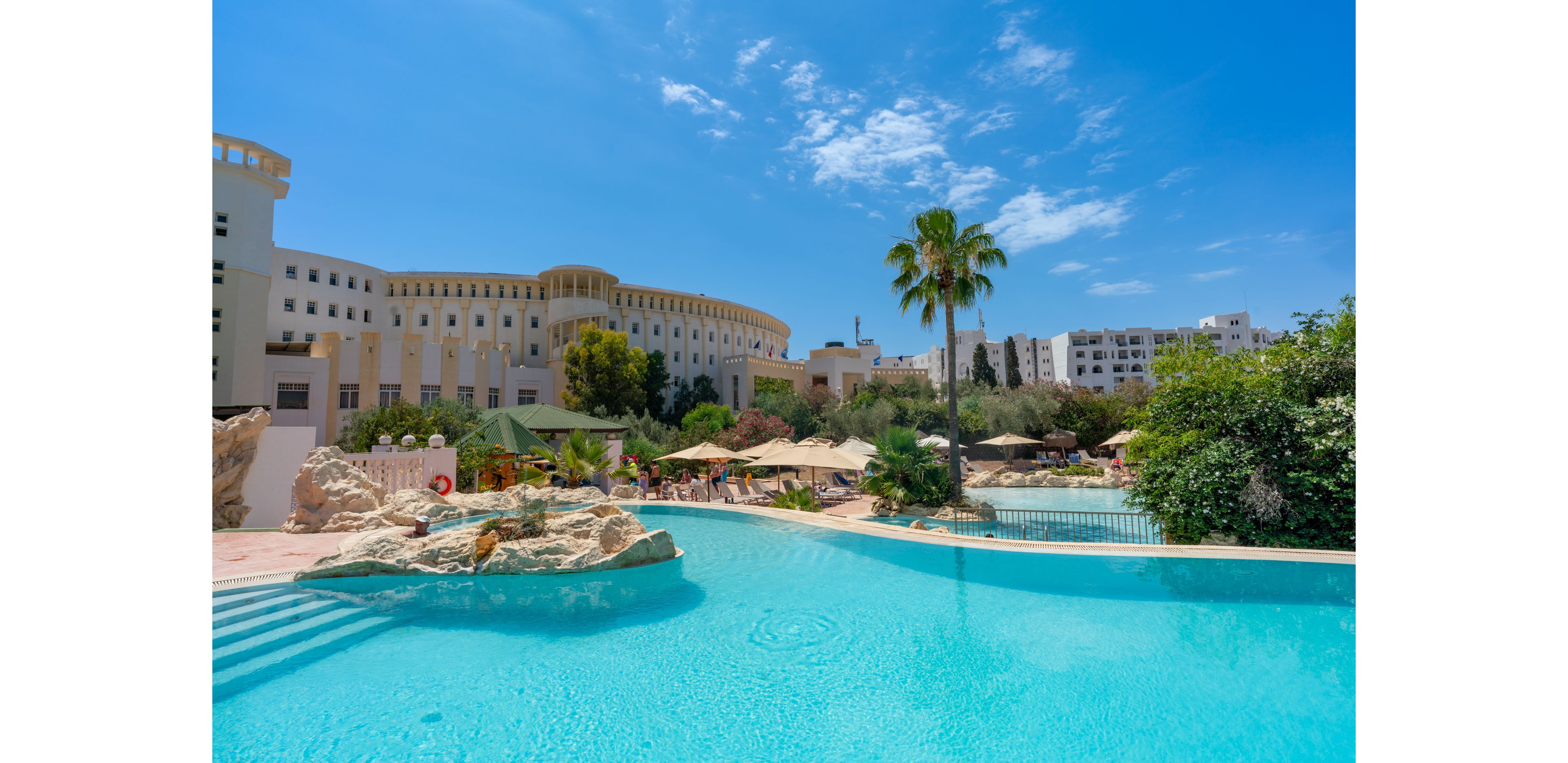 A large turquoise swimming pool stretches across the foreground of a sunny resort. Guests relax under umbrellas and shade structures beside the water, surrounded by lush greenery and palm trees. In the background, a multi-story hotel building with arched windows rises against a bright blue sky. The scene conveys a warm, inviting vacation atmosphere with clear weather and calm water.