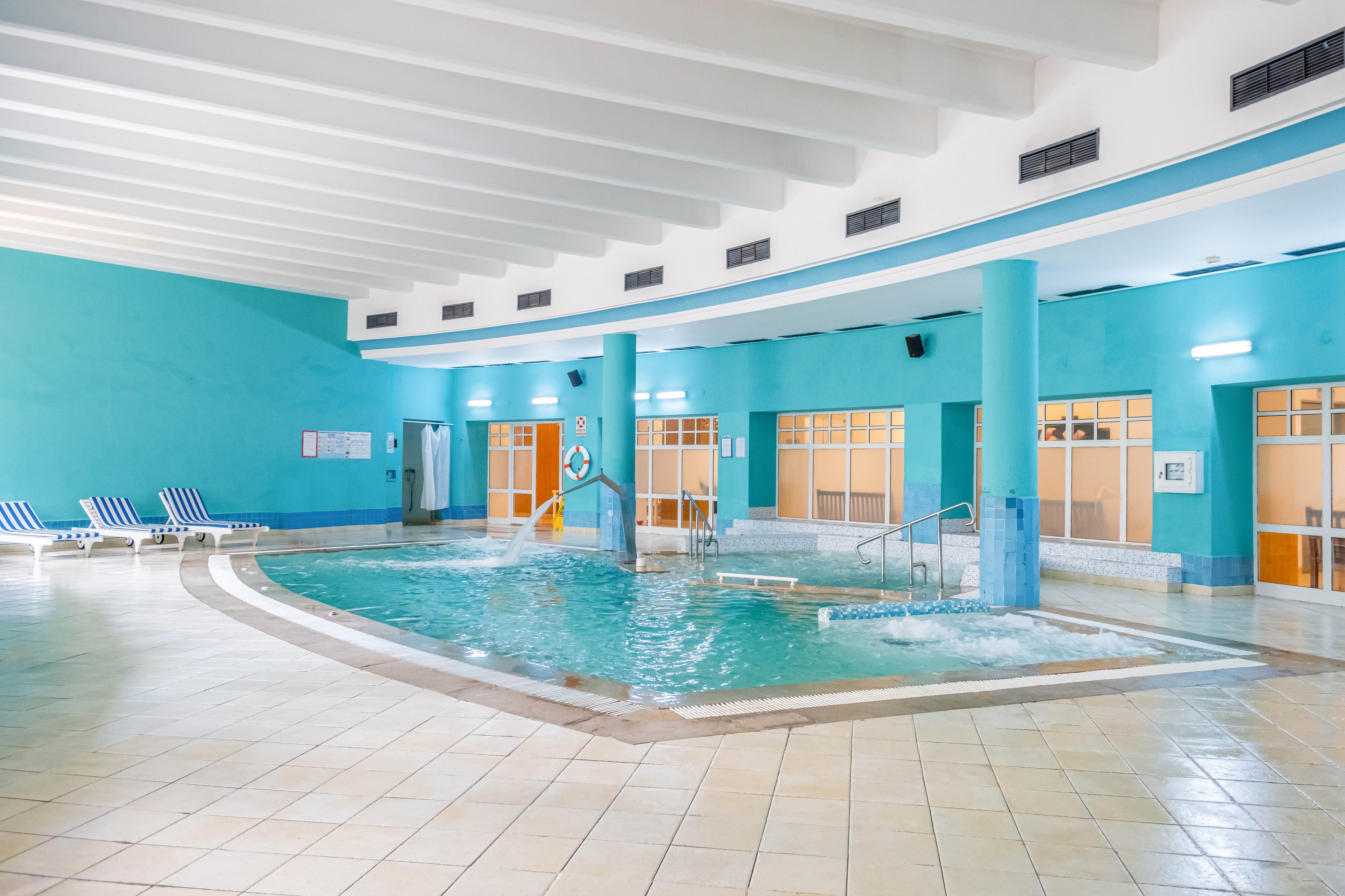 An indoor spa pool is featured in a spacious, brightly lit room with turquoise walls and tiled floors. The pool includes metal handrails, water jets, and a shallow entry area, suggesting a relaxing hydrotherapy environment. Several striped lounge chairs line the far wall, while large windows and doors provide natural light. The clean architectural lines and soft color palette create a calm, modern wellness atmosphere.