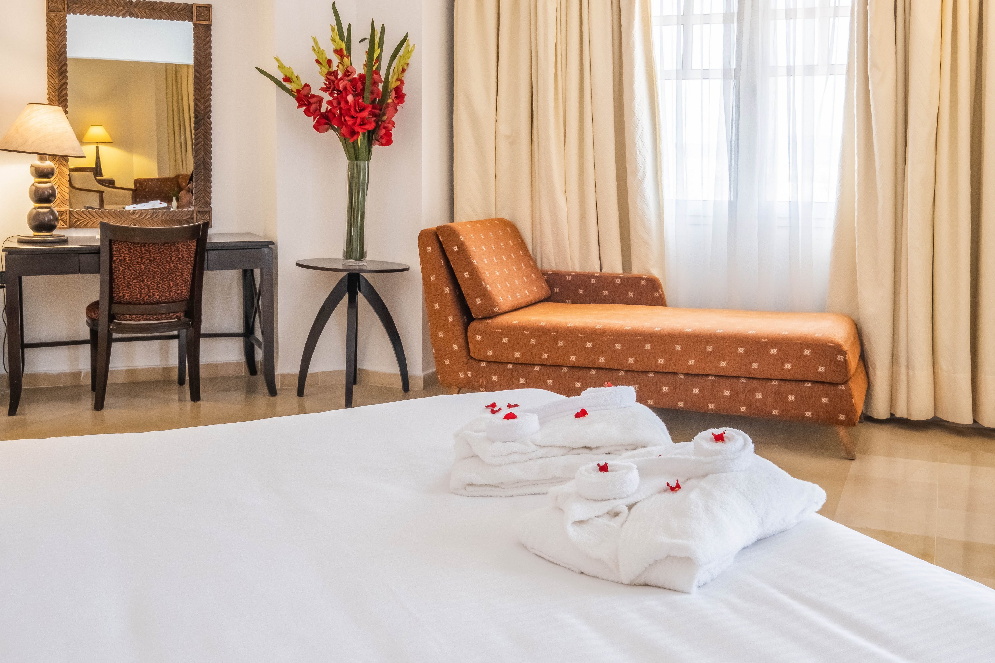 A neatly made hotel bed with folded white bathrobes is arranged in the foreground of a bright bedroom. In the background, a cushioned chaise lounge sits beside large curtained windows, allowing soft daylight to fill the room. A dark wooden desk with a mirror, lamp, and a vase of red flowers adds a touch of sophistication. The overall scene suggests a comfortable, welcoming space for relaxation and travel accommodation.