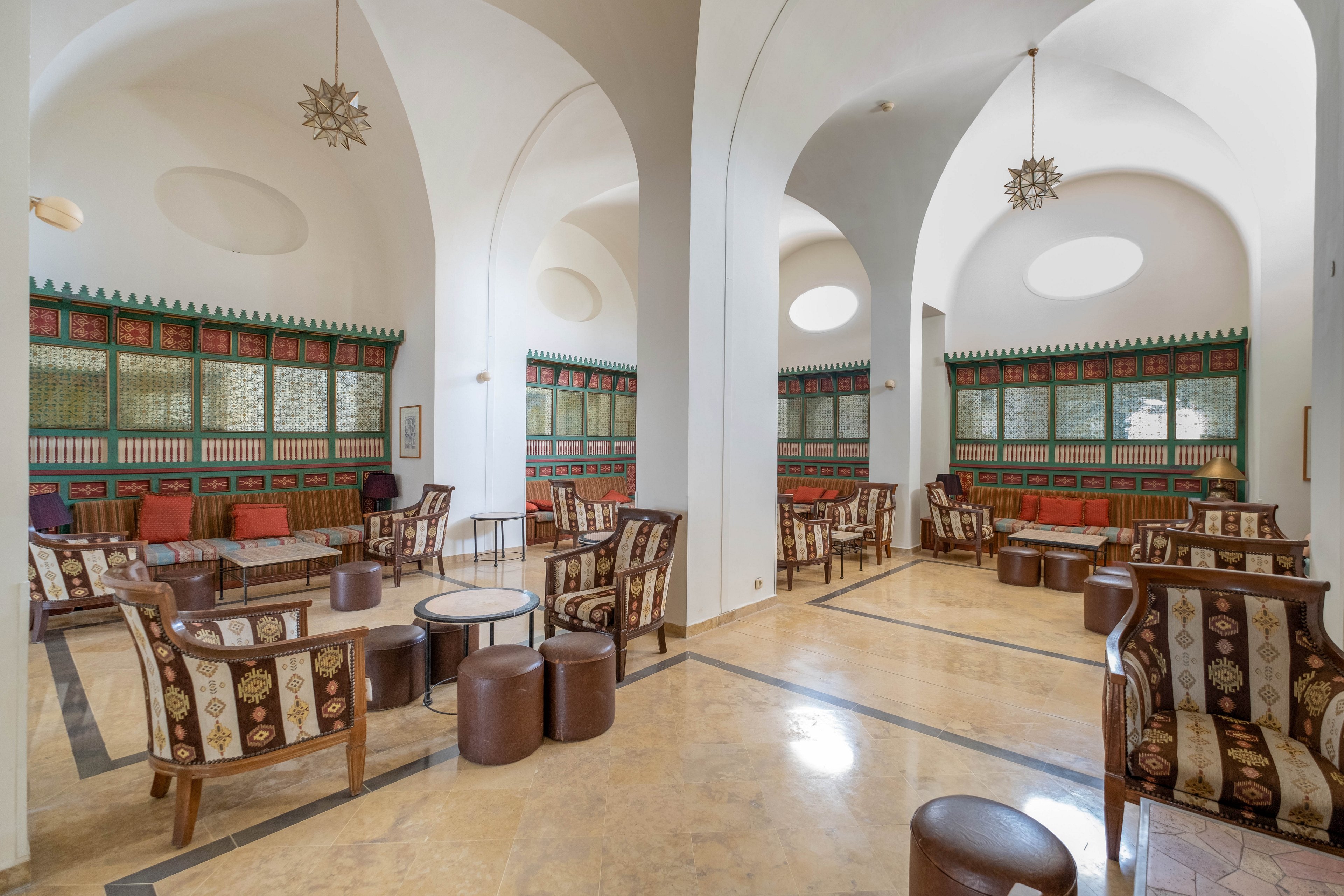 A spacious lounge features high white vaulted ceilings and large arches creating an airy, open atmosphere. Numerous upholstered armchairs and round tables are arranged in conversational clusters across a polished stone floor. The walls are decorated with colorful geometric panels and a repeating pattern of small arches, adding a traditional decorative touch. Ornate pendant lights hang from the ceiling, and natural light filters through circular skylights, enhancing the serene and refined setting.