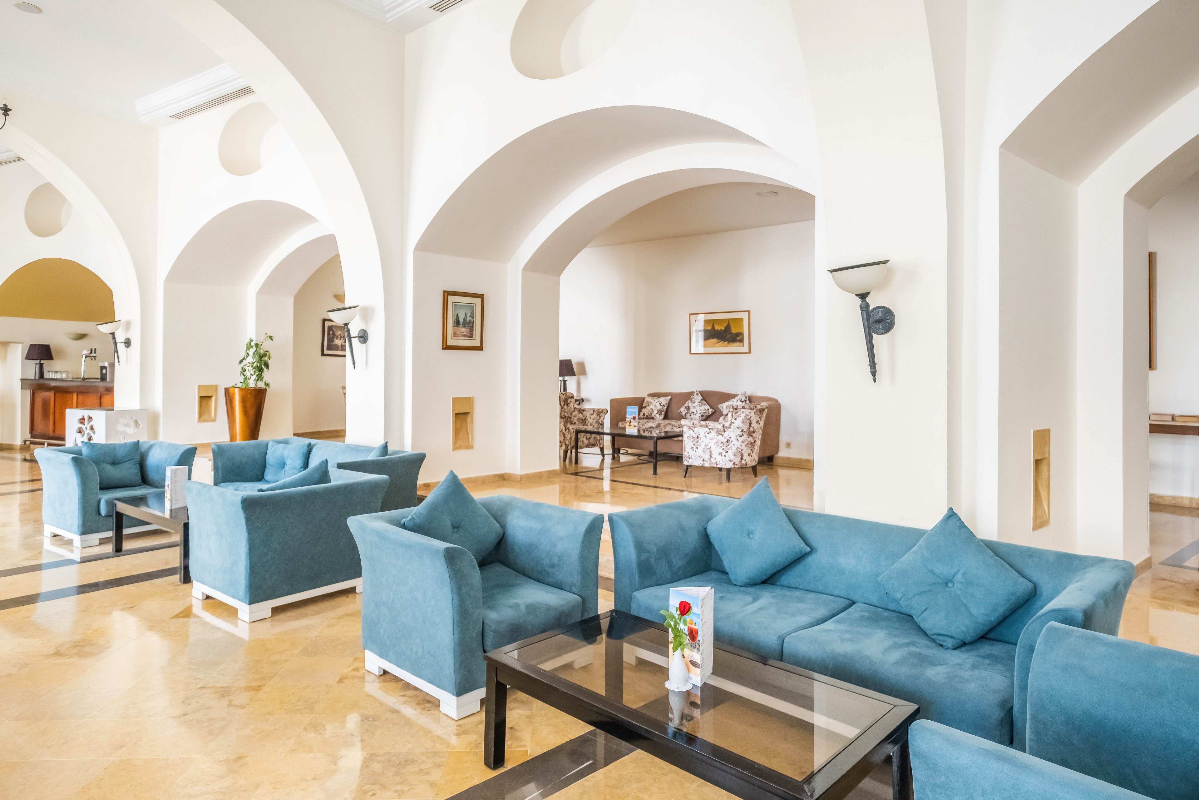 A spacious hotel lobby features multiple blue upholstered sofas arranged around glass coffee tables. The room has high arched ceilings, cream-colored walls, and polished marble floors that reflect the natural light. Framed artwork and decorative plants add visual interest to the otherwise minimalist interior. In the background, additional seating areas and a reception or bar counter contribute to the welcoming atmosphere.