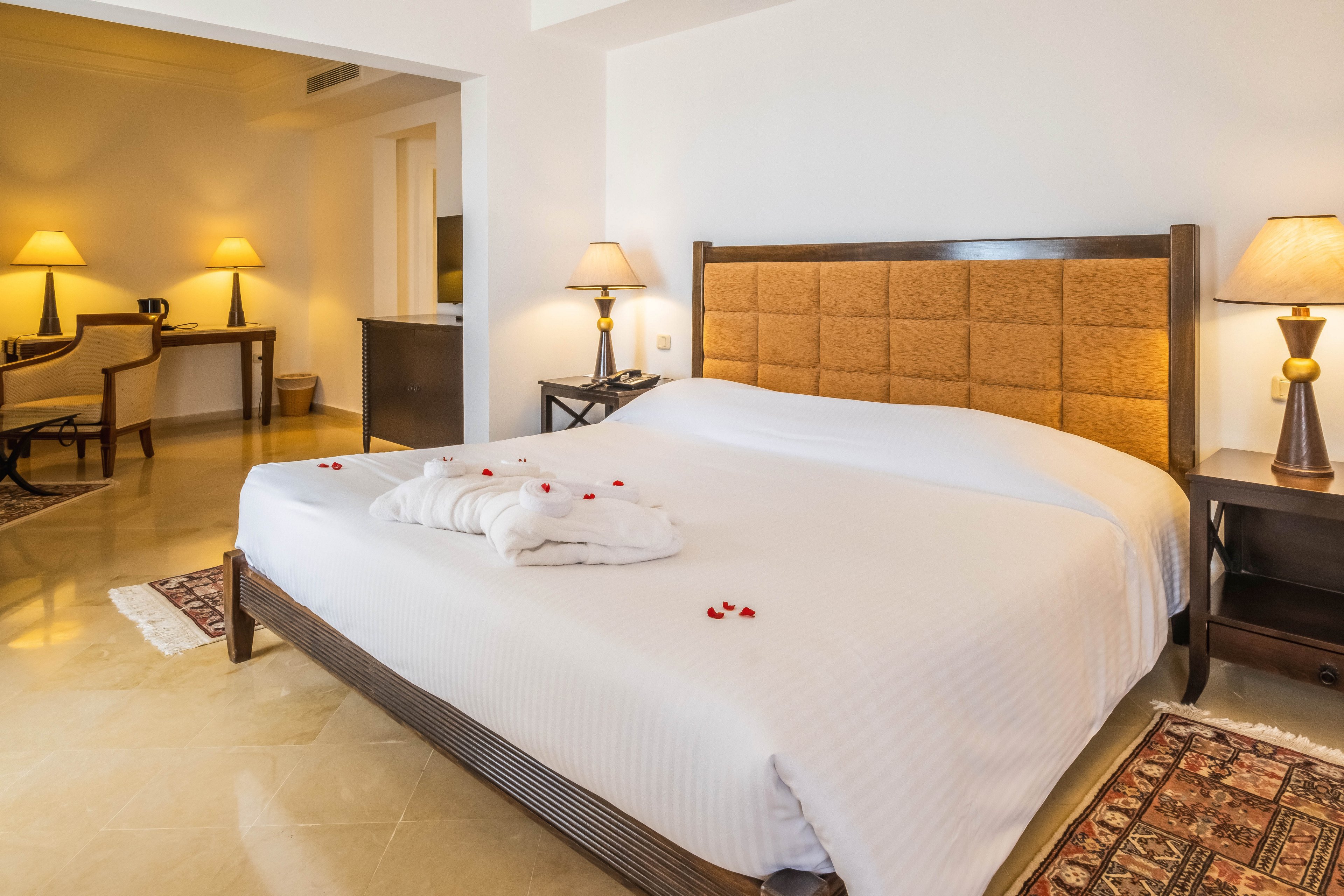 A neatly made bed with a padded headboard is the focal point of this cozy hotel bedroom. White linens and a folded bathrobe with scattered flower petals create a welcoming, romantic touch. Warm table lamps on bedside tables and in the adjoining sitting area cast a soft glow across the room. Neutral walls, tiled floors, and patterned rugs contribute to a clean, modern yet comfortable atmosphere.