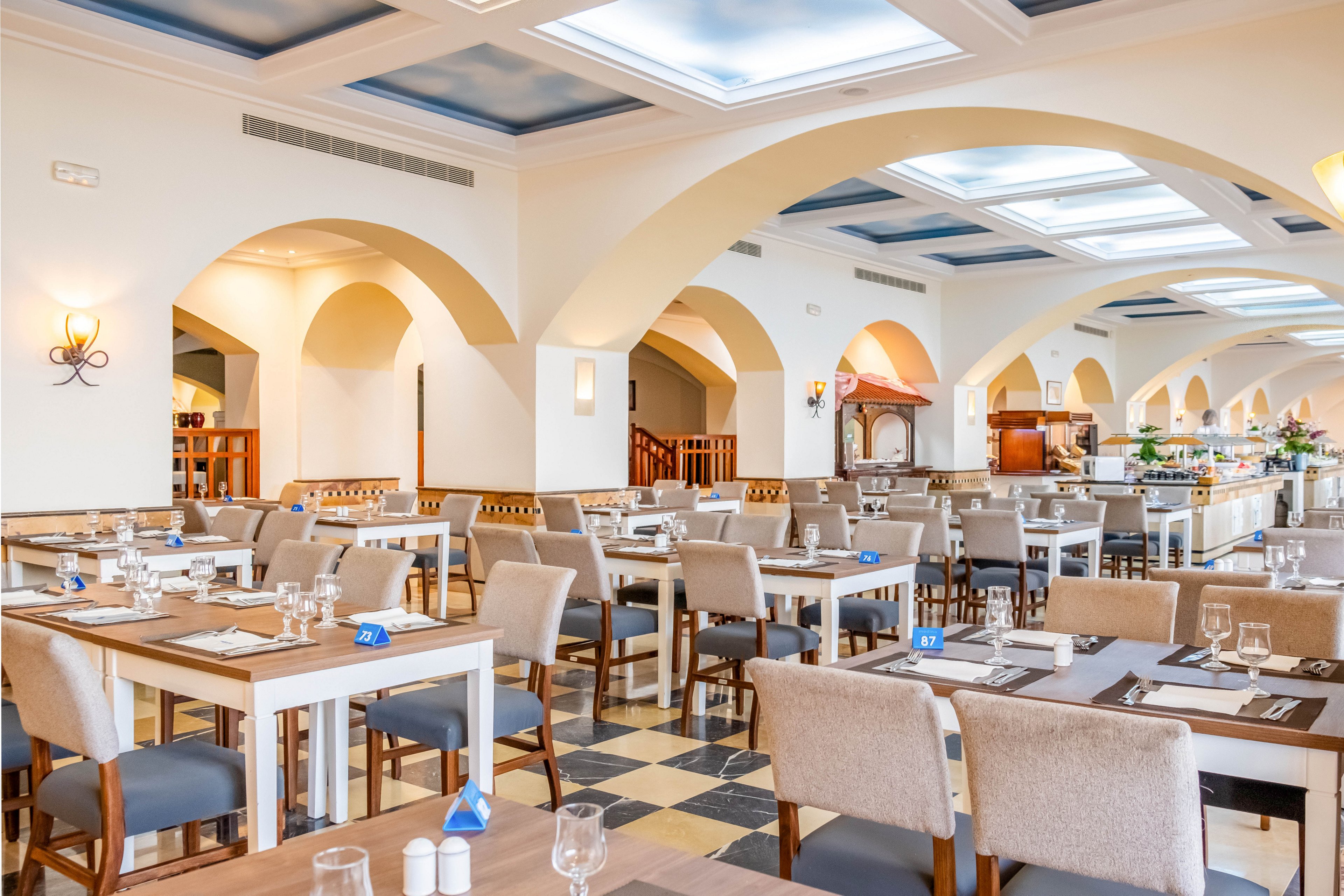 Spacious restaurant dining room with neatly arranged tables and chairs. The setting features arched ceilings, large skylights, and warm wall sconces that create a bright, inviting atmosphere. Tables are set with glassware, cutlery, napkins, and small table number stands. The background shows a buffet or service area and architectural details in neutral tones. The overall scene suggests a clean, contemporary dining experience in a hotel or resort.
