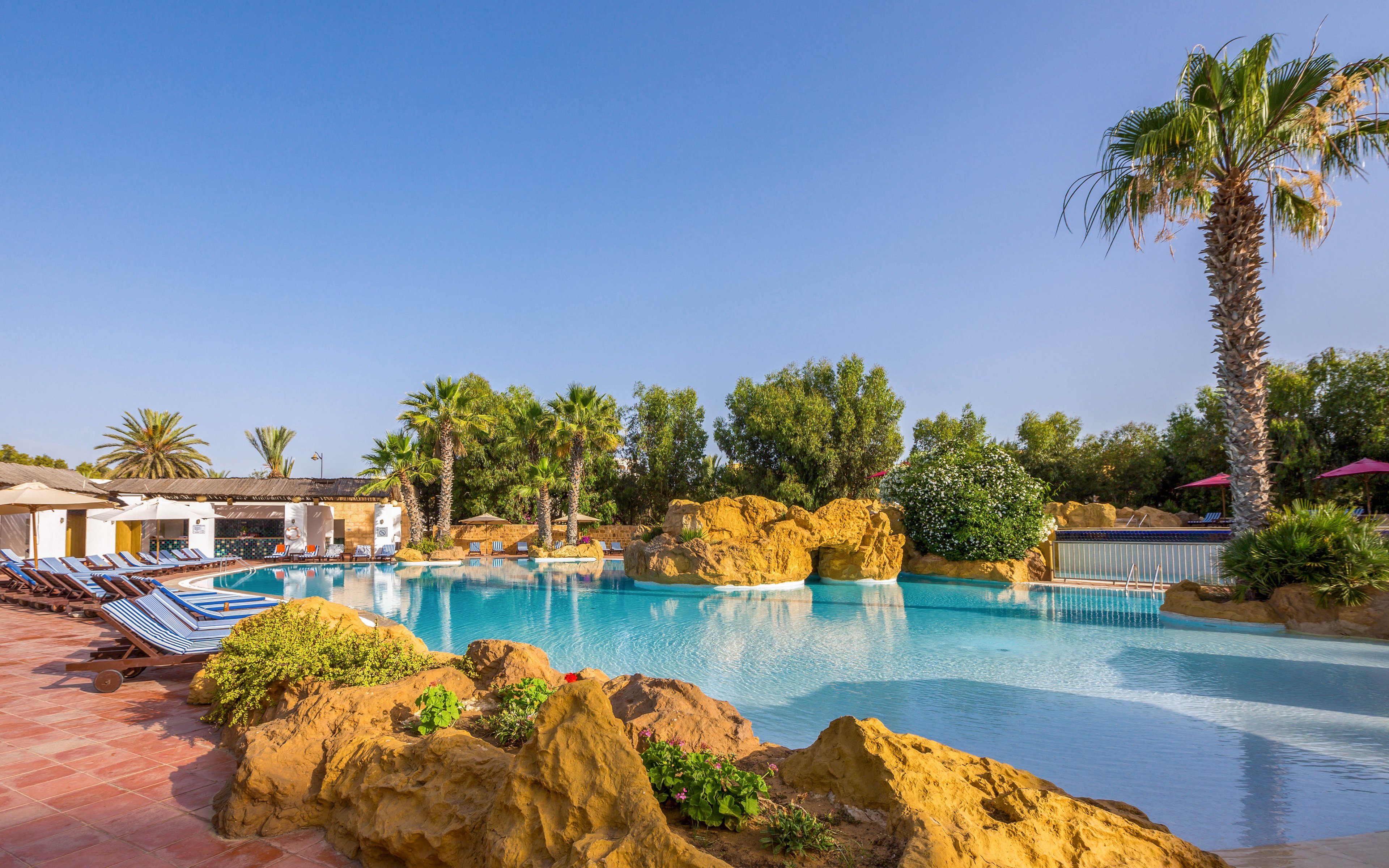 A large resort-style swimming pool is surrounded by palm trees and rocky landscaping under a clear blue sky. Sun loungers line the tiled poolside area, suggesting a relaxing vacation setting. The water is calm and inviting, reflecting the surrounding greenery and bright daylight. Buildings and shaded areas in the background indicate amenities for guests enjoying the warm weather.