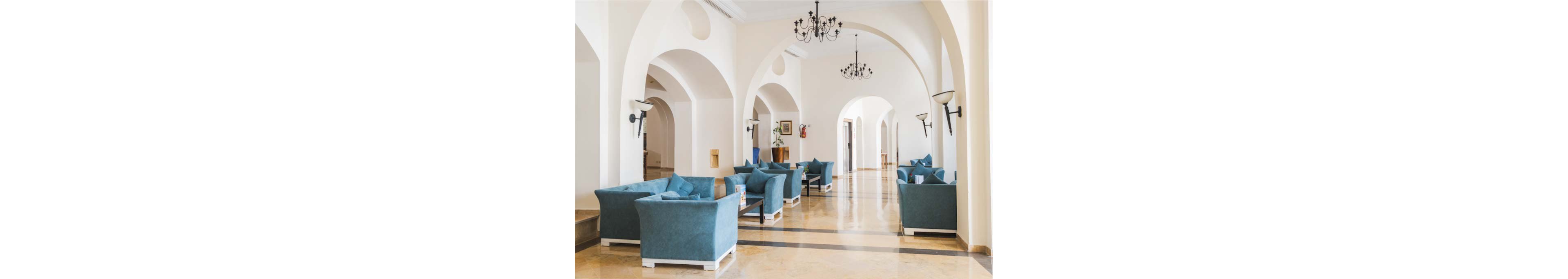 Spacious hotel lobby with multiple blue armchairs arranged in small seating groups. The interior features high white walls, large arches, and polished marble floors. Black wrought-iron chandeliers and wall sconces provide classic decorative lighting. Natural light floods the corridor, creating an airy and welcoming atmosphere. The space appears quiet and ready for guests to relax or wait comfortably.