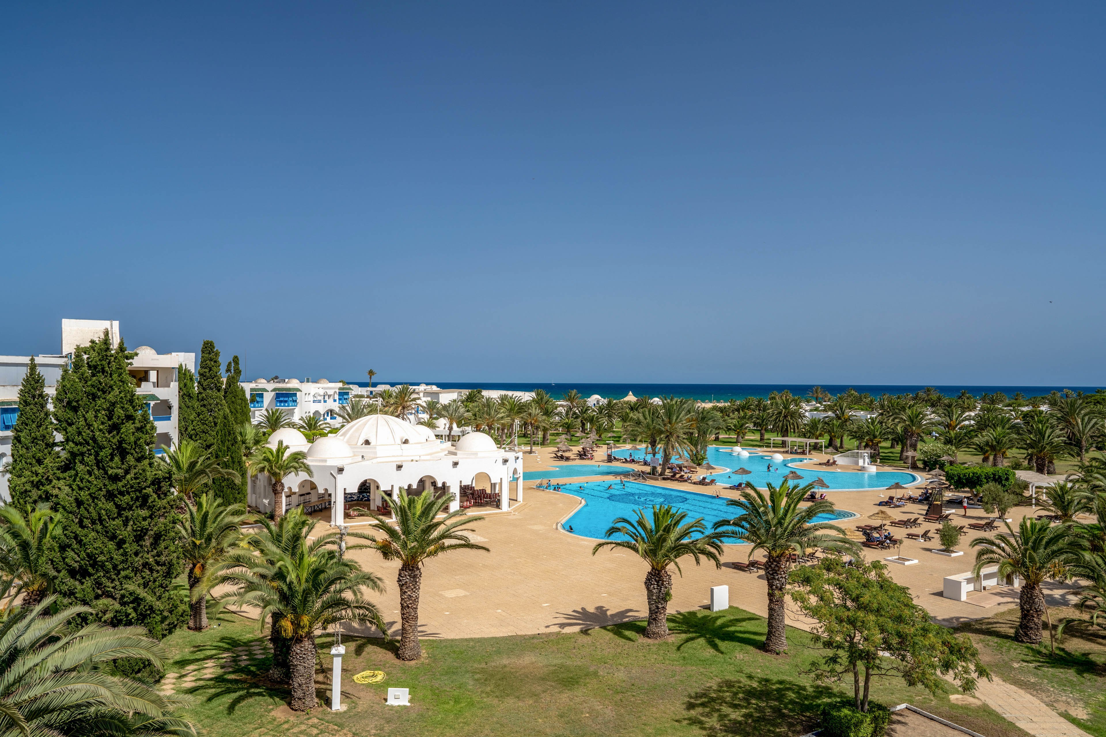 A large seaside resort features multiple blue swimming pools surrounded by palm trees and landscaped gardens. White buildings with domed roofs and Mediterranean-style architecture frame the pool area. Sun loungers and umbrellas are arranged around the pools, suggesting a relaxing vacation setting. In the distance, the deep blue sea meets a clear cloudless sky, enhancing the tranquil, sunny atmosphere.