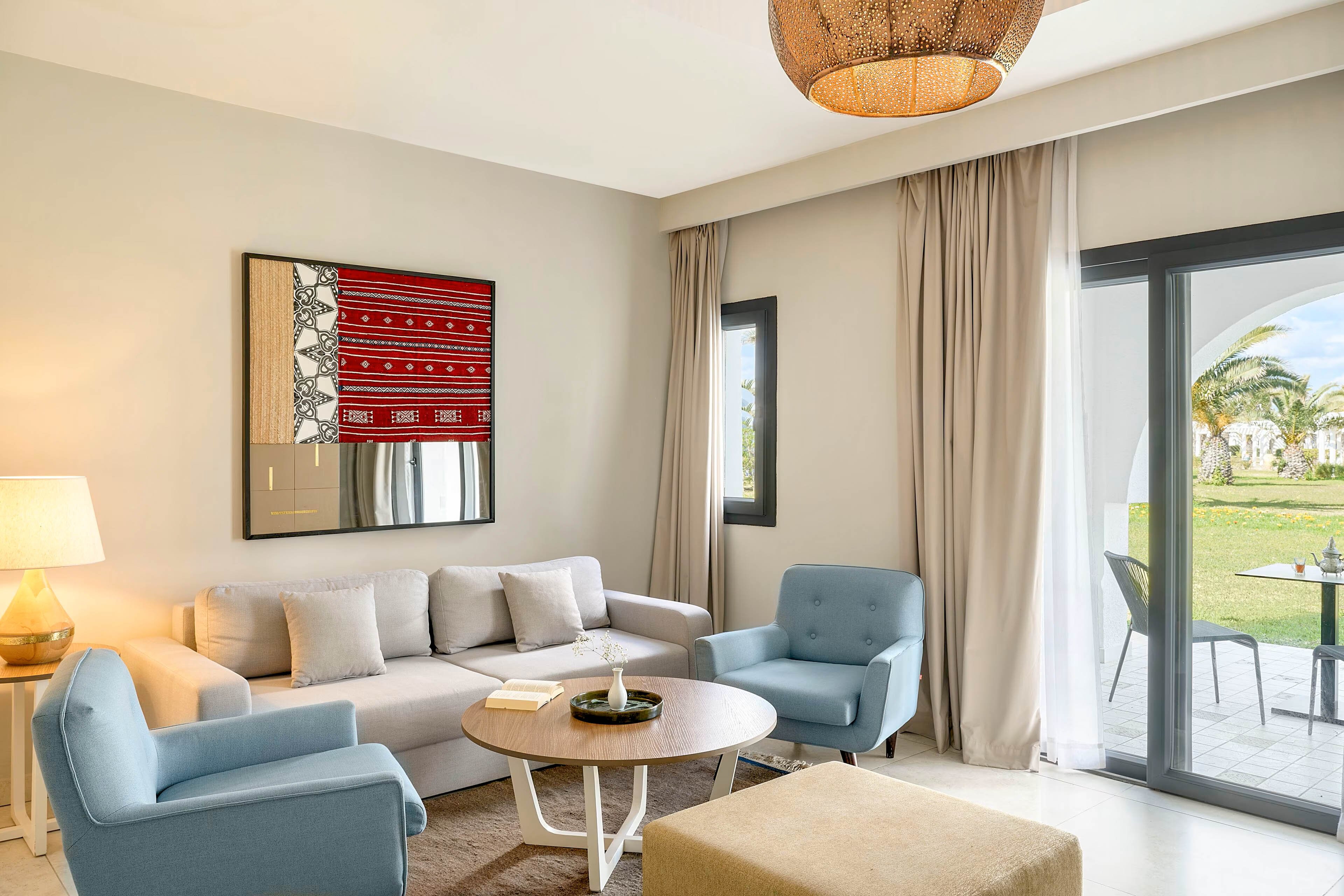 A contemporary living room features a light gray sofa paired with blue armchairs and a round coffee table. Neutral walls and beige curtains frame large glass doors that open to a sunny garden and patio area. A woven ceiling light and framed wall art add warmth and texture to the minimalist decor. Soft natural light fills the space, creating an inviting and relaxed atmosphere.