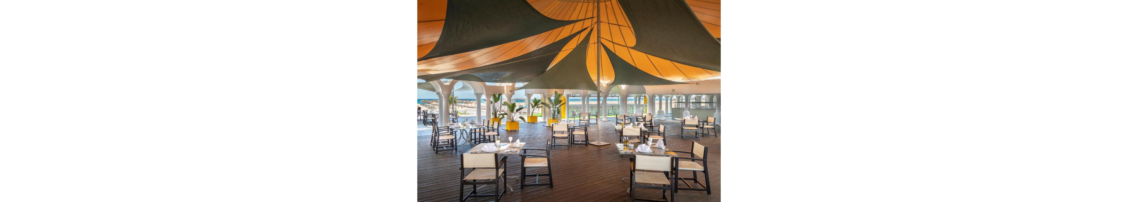 An open-air restaurant features wooden flooring and light-colored chairs arranged around small tables. A large, sculptural canopy in green and yellow shades provides shade over the dining area. Arched openings frame views of the beach and ocean in the background. Potted plants in bright yellow containers add a tropical touch to the relaxed seaside setting.