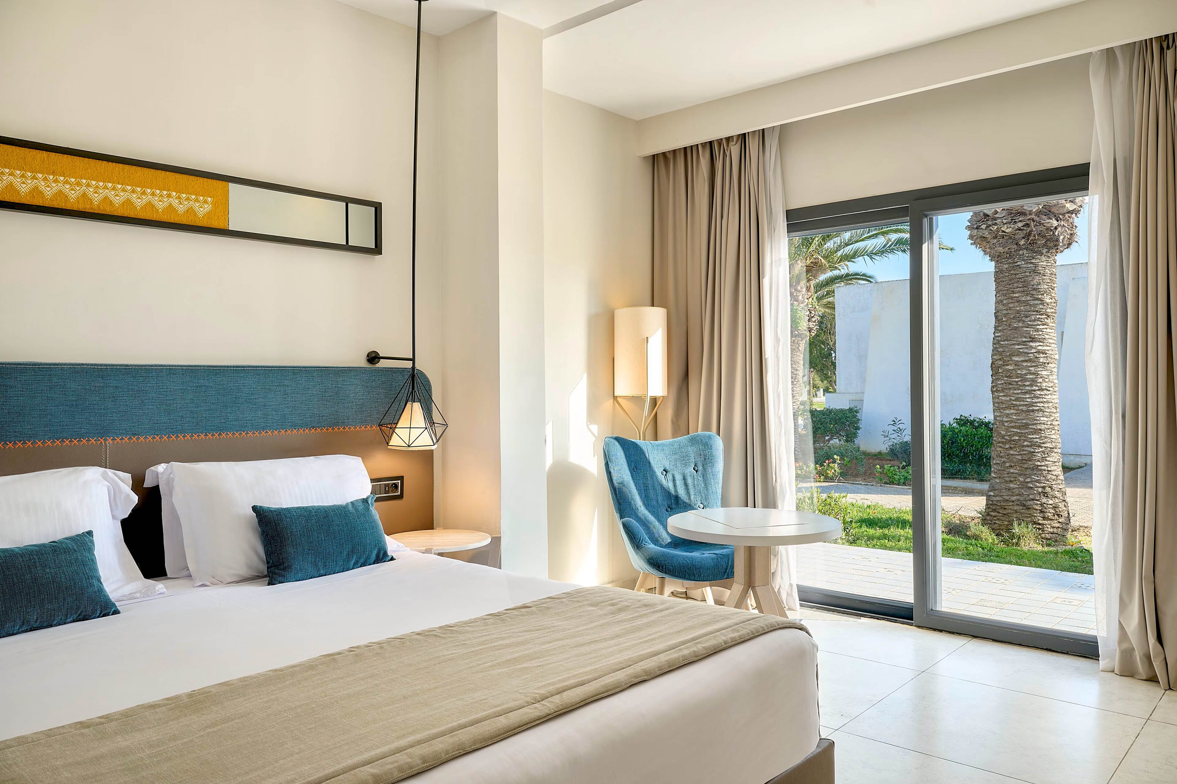A contemporary hotel room features a neatly made double bed with white linens and a beige throw. The space includes a blue upholstered headboard, a matching blue accent chair, and a small round table near the window. Large sliding glass doors with light curtains open to a sunny garden area with palm trees and white buildings. Soft neutral tones and natural light create a calm, inviting atmosphere suitable for relaxation or vacation stays.