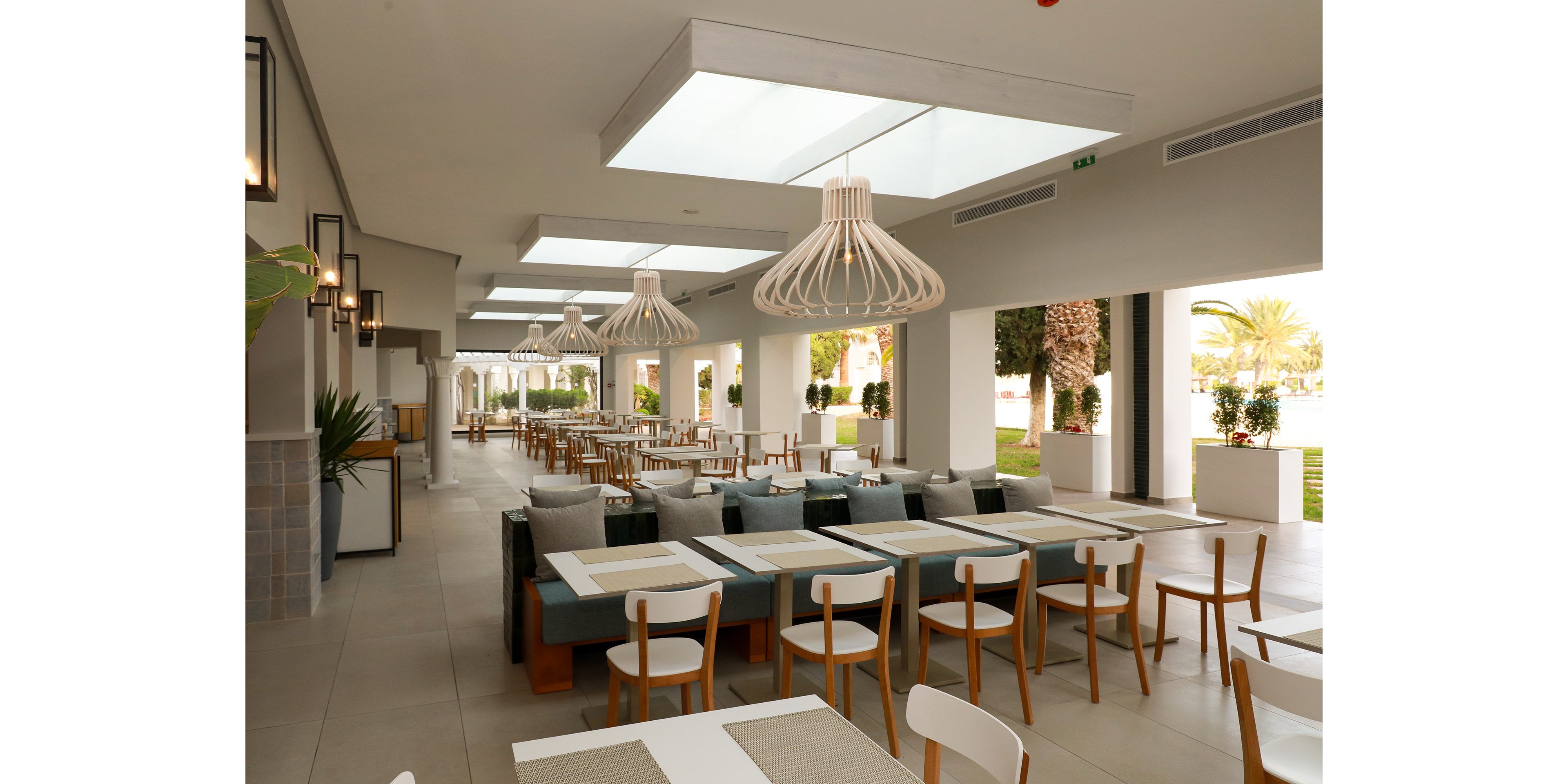 A spacious modern restaurant interior features long tables with wooden chairs and cushioned bench seating. Large geometric pendant lights hang from the ceiling, illuminating the neutral-toned space. The open-air design leads to an outdoor area with palm trees and bright natural light. Clean lines, minimal decor, and soft colors create a relaxed, contemporary dining atmosphere.