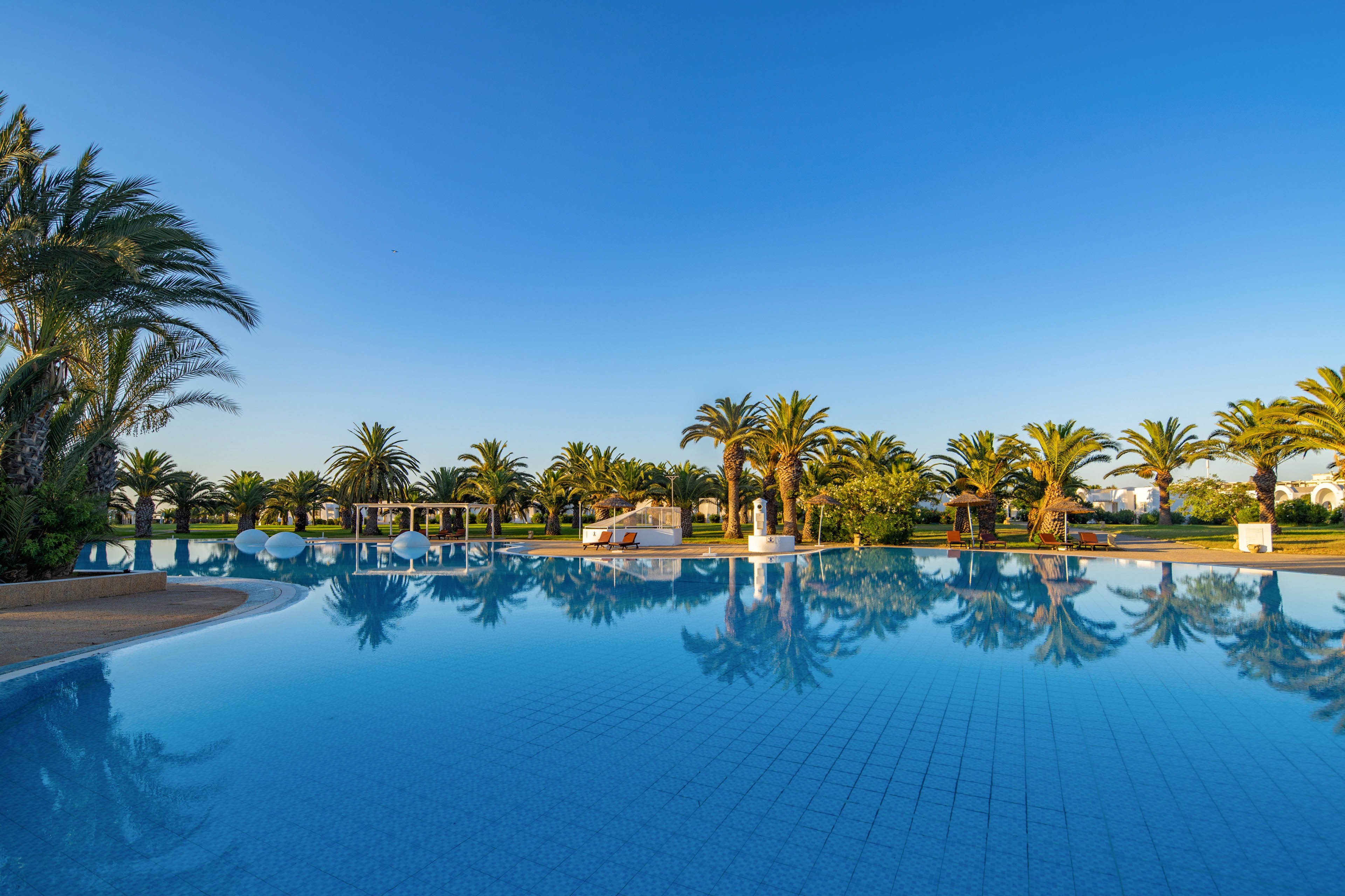 A large, tranquil swimming pool reflects palm trees and a clear blue sky at a tropical resort. The water is calm, creating mirror-like reflections of the surrounding greenery and structures. White lounge areas and small buildings line the far edge of the pool, suggesting a relaxing vacation setting. The bright morning light and cloudless sky emphasize a serene, warm atmosphere.
