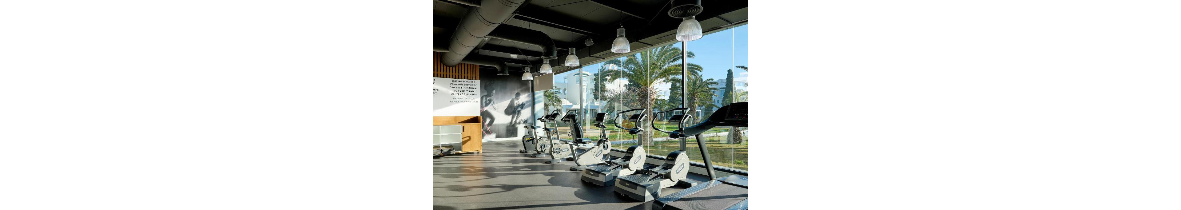 A bright, modern fitness room features several stationary bikes and treadmills neatly arranged in front of large floor-to-ceiling windows. Outside, palm trees and a landscaped area create a resort-like atmosphere visible from the gym. Overhead industrial-style lighting and exposed ductwork contribute to a contemporary interior design. A wall graphic of an athlete and a text panel add a motivational sports theme to the space.