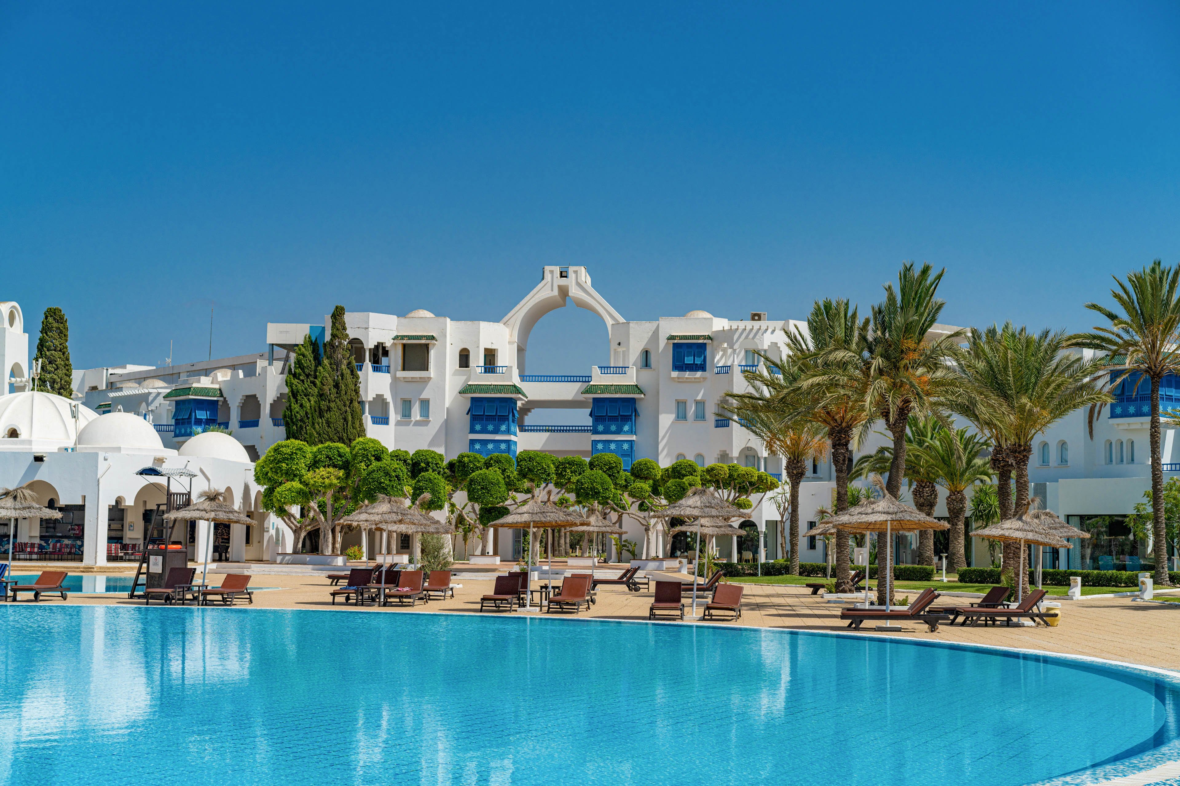 A large white resort hotel stands behind a bright blue swimming pool under a clear sky. Sun loungers and straw parasols line the poolside terrace, suggesting a relaxing vacation setting. Tall palm trees and manicured greenery frame the scene, adding a tropical atmosphere. The architecture features arches and domes, giving the property a distinctive Mediterranean or North African style.