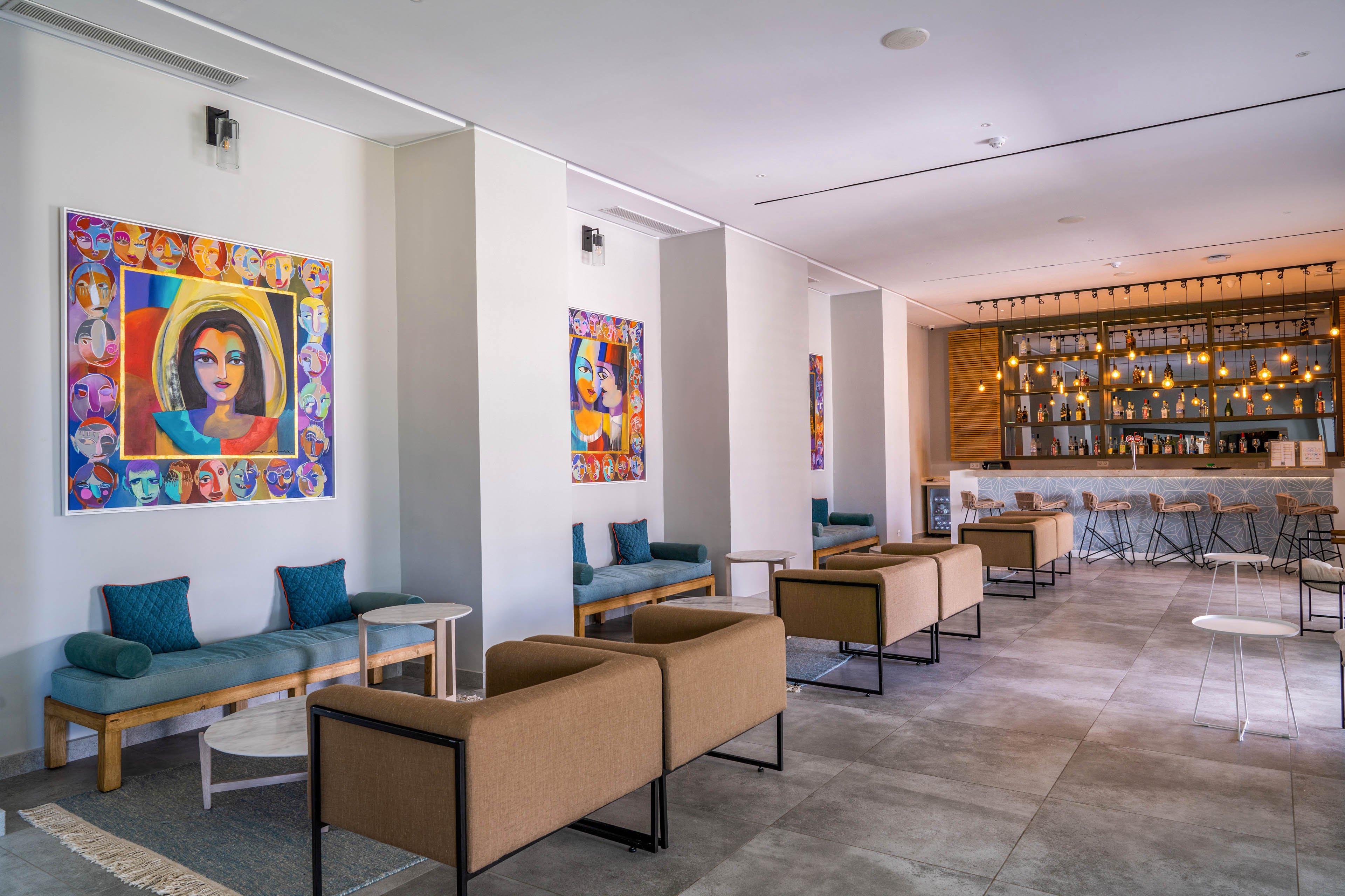 A contemporary hotel lobby features a series of minimalist seating areas with cushioned benches and armchairs. Colorful abstract artwork hangs on the walls, adding vibrant accents to the clean white interior. In the background, a stylish bar with high stools and warm pendant lighting creates an inviting atmosphere. The space appears bright, open, and designed for casual socializing or relaxation.