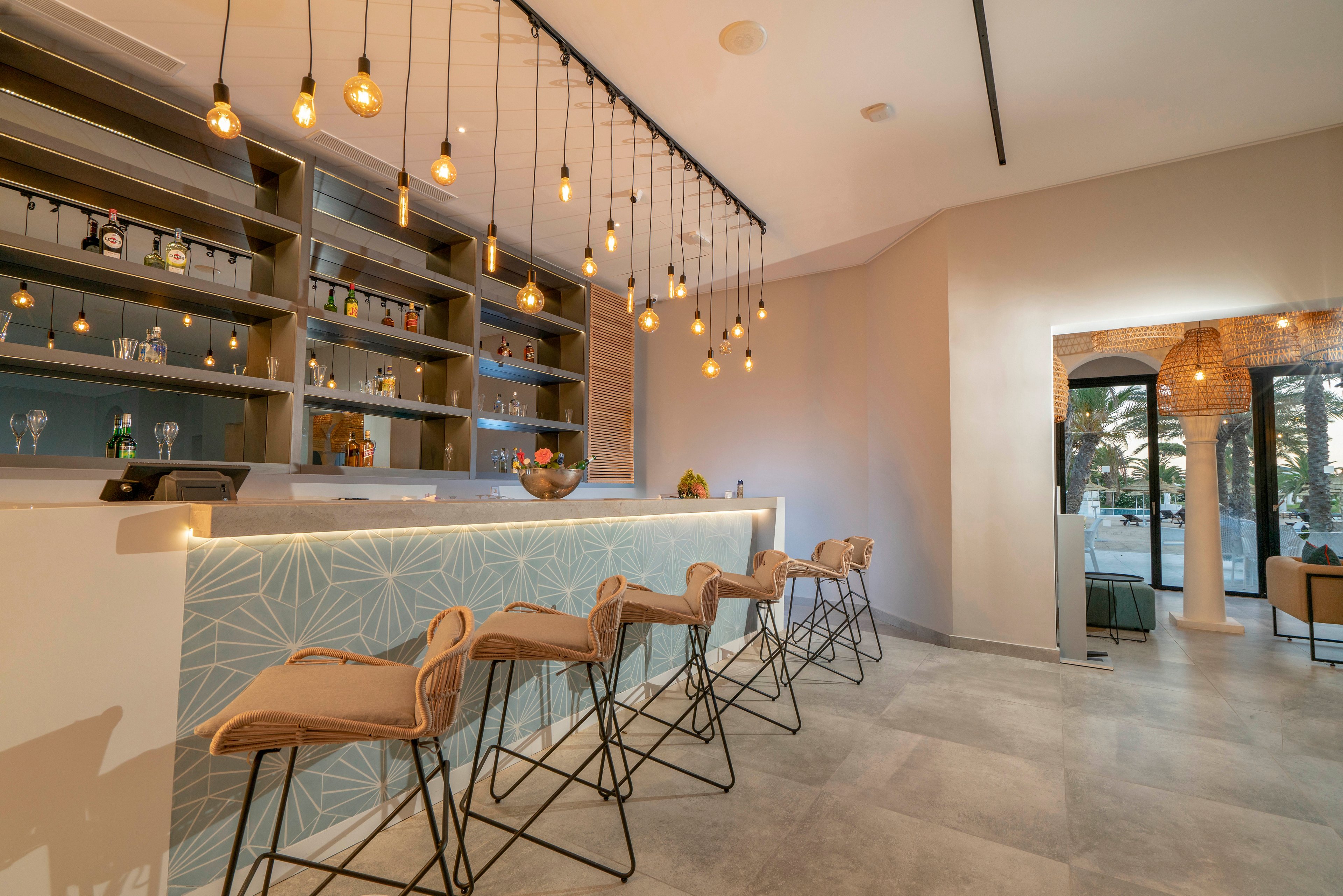 A contemporary bar interior features a long counter with patterned front paneling and a row of high stools. Warm pendant lights hang from the ceiling, casting a soft glow over the shelves of bottles and glassware. The space has a minimalist design with neutral tones, clean lines, and polished flooring. Large windows and an open doorway reveal an adjacent lounge area and outdoor greenery, enhancing the airy, inviting atmosphere.