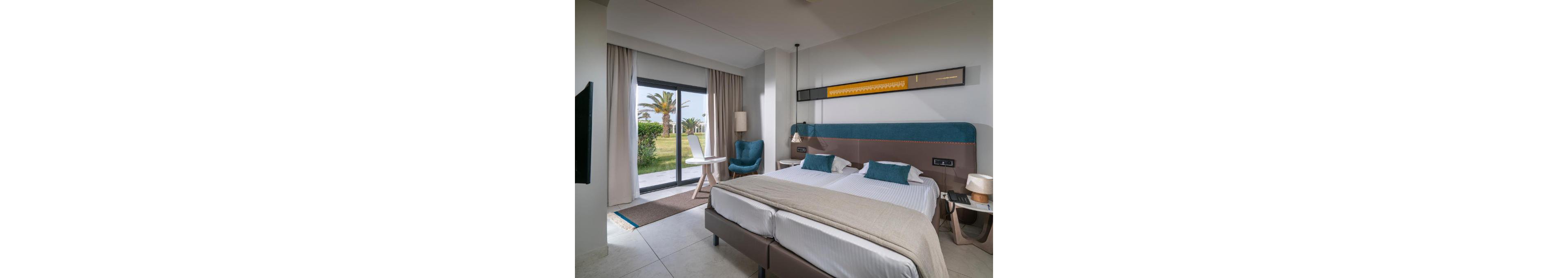 A contemporary hotel room features a large double bed with neutral bedding and a teal upholstered headboard. The space includes a small round table with two chairs near a sliding glass door that opens to a green garden with palm trees. Soft natural light enters through sheer curtains, complemented by a floor lamp and bedside lamps. Minimalist decor and clean lines create a calm, inviting atmosphere suitable for relaxation or vacation stays.