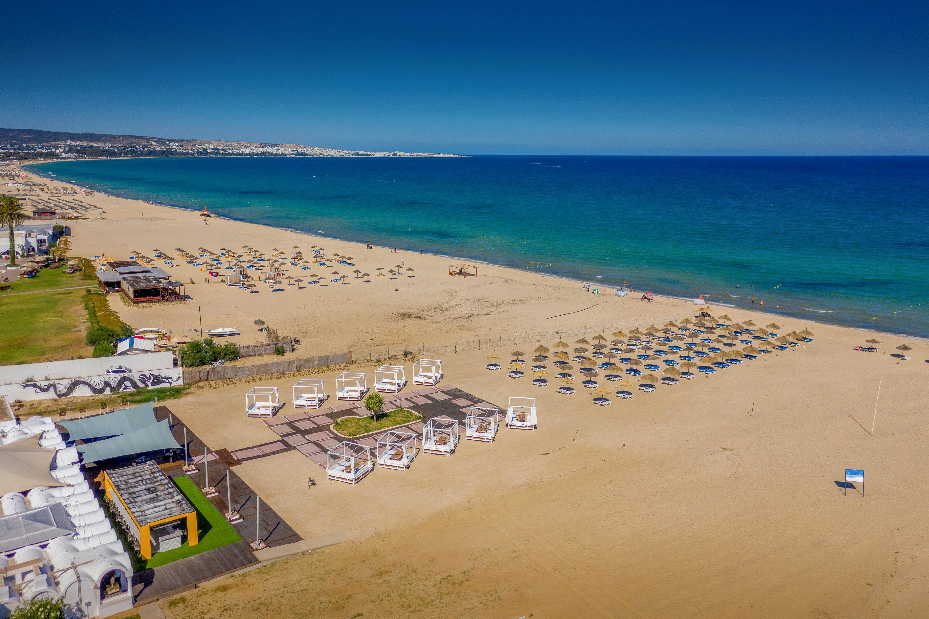 A wide sandy beach stretches along a bright blue coastline under a clear sky. Numerous sun loungers and umbrellas are neatly arranged in rows near the shoreline. A section of white cabanas and a nearby beach club structure occupy the foreground. The calm sea and distant coastline create a relaxed holiday atmosphere. The scene suggests a well-organized seaside resort prepared for visitors.