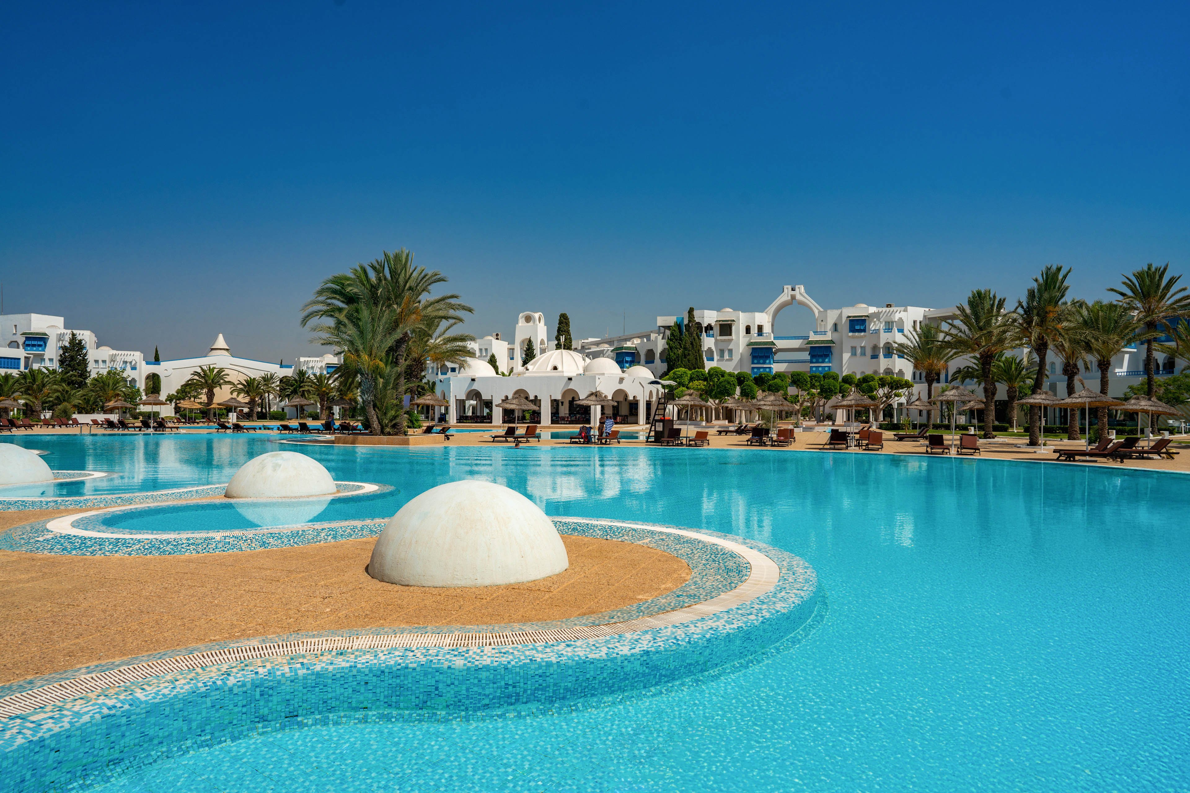 A large turquoise swimming pool dominates the foreground of a luxury resort. Curved sand-colored islands and white domed structures create an artistic design within the water. In the background, white buildings with arches and balconies line the poolside, surrounded by tall palm trees. Sun loungers and umbrellas suggest a relaxing holiday atmosphere under a clear blue sky.