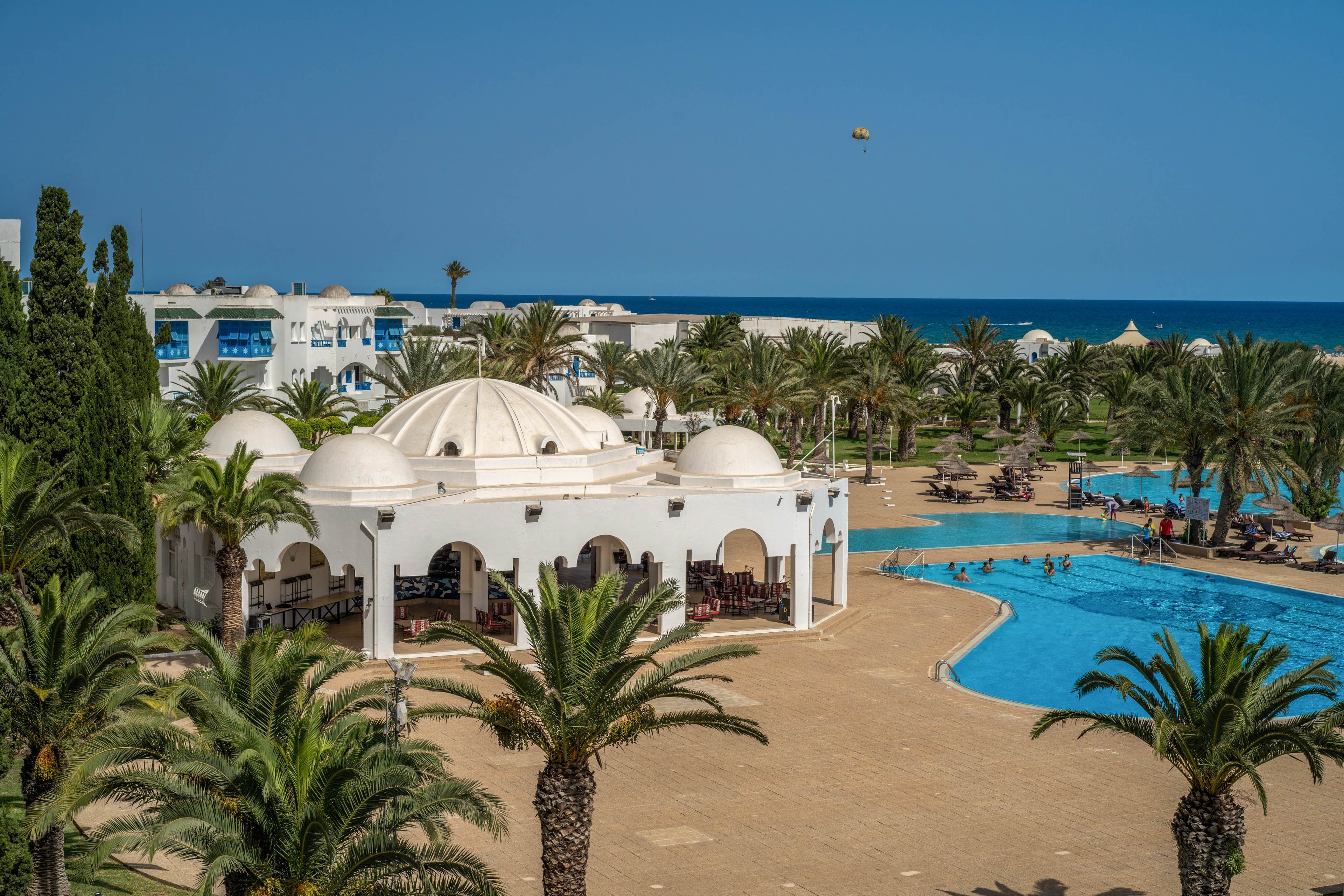 A coastal resort features a white domed pavilion beside a large outdoor swimming pool. Palm trees line the pool deck and extend toward the horizon, creating a lush oasis atmosphere. White low-rise buildings with arches and balconies surround the area, while the blue sea is visible in the background under a clear sky. The scene suggests a warm, sunny vacation destination focused on relaxation and leisure by the water.