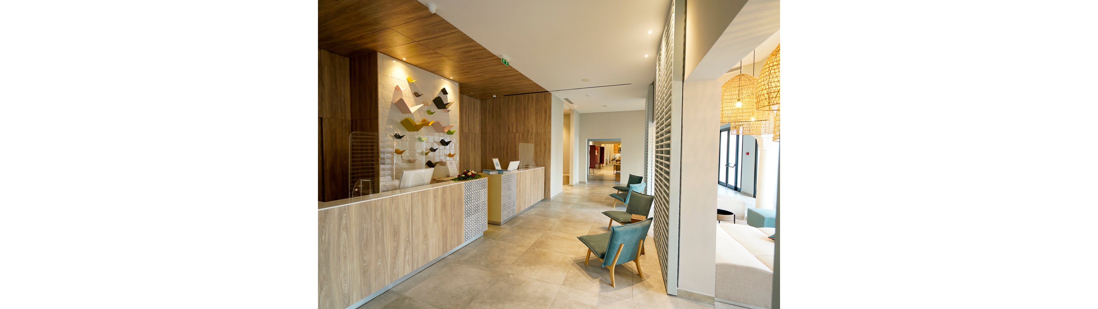 A contemporary hotel reception area features a long wooden front desk with minimalist decor. The space includes a wall art installation behind the counter and warm wood paneling on the walls and ceiling. Comfortable armchairs line the corridor, leading toward a bright lounge area with large windows. Pendant lights with woven shades add a cozy ambiance to the clean, modern interior.