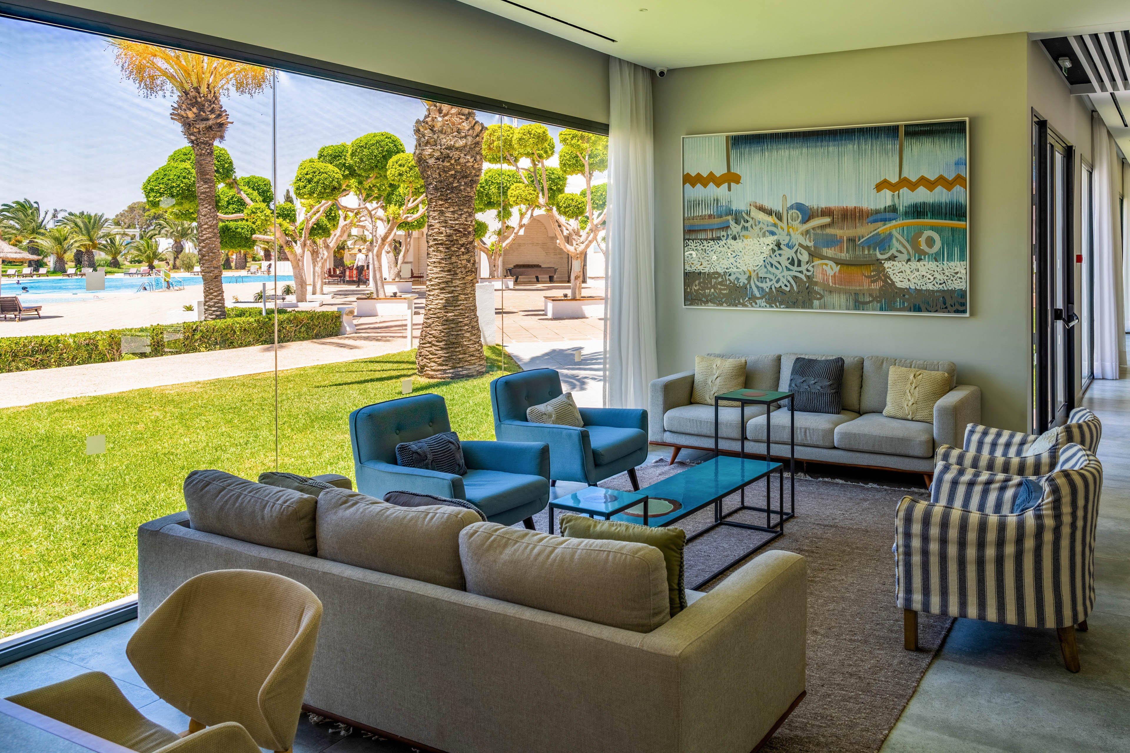 A contemporary lounge area features comfortable sofas and armchairs arranged around low coffee tables. Floor-to-ceiling windows open onto a lush green lawn with sculpted trees and a bright outdoor pool area. Neutral-toned seating is accented by blue chairs and a striped armchair, while a large framed artwork hangs on the wall. The scene suggests a relaxed, upscale resort environment with abundant natural light.