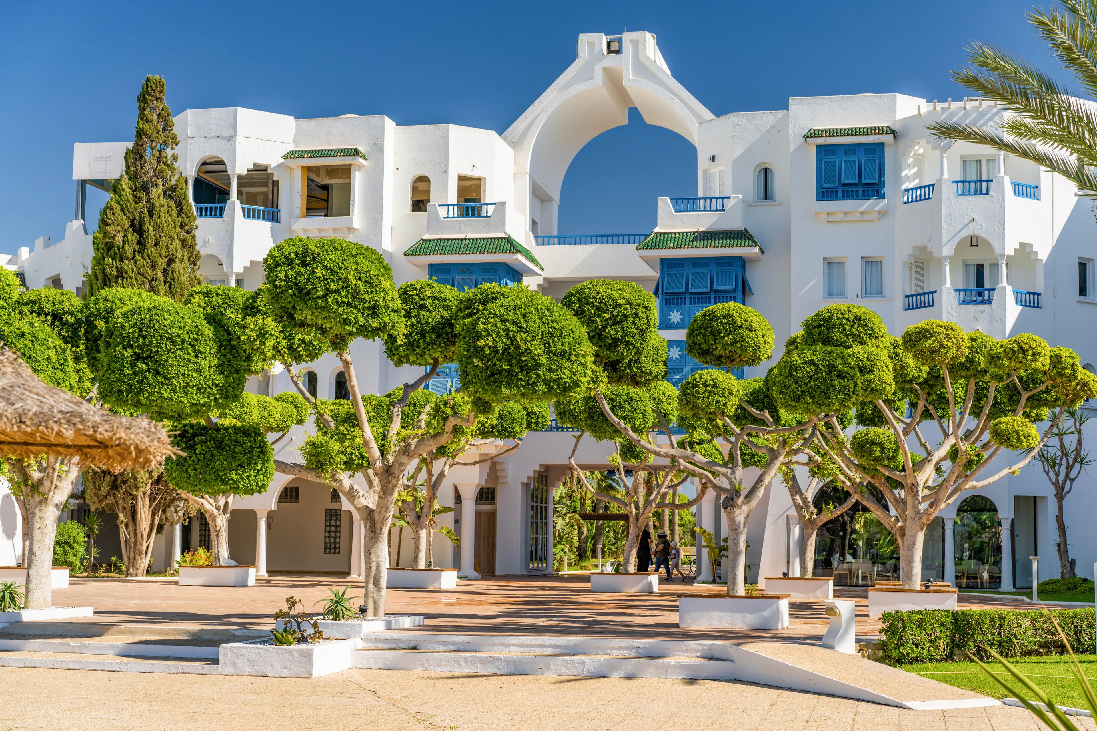 A bright white Mediterranean-style building stands under a clear blue sky. The architecture features arches, balconies, and vivid blue window shutters. In the foreground, neatly trimmed green trees and landscaped pathways create a welcoming courtyard. A few people walk near the entrance, adding a sense of scale and calm resort atmosphere.