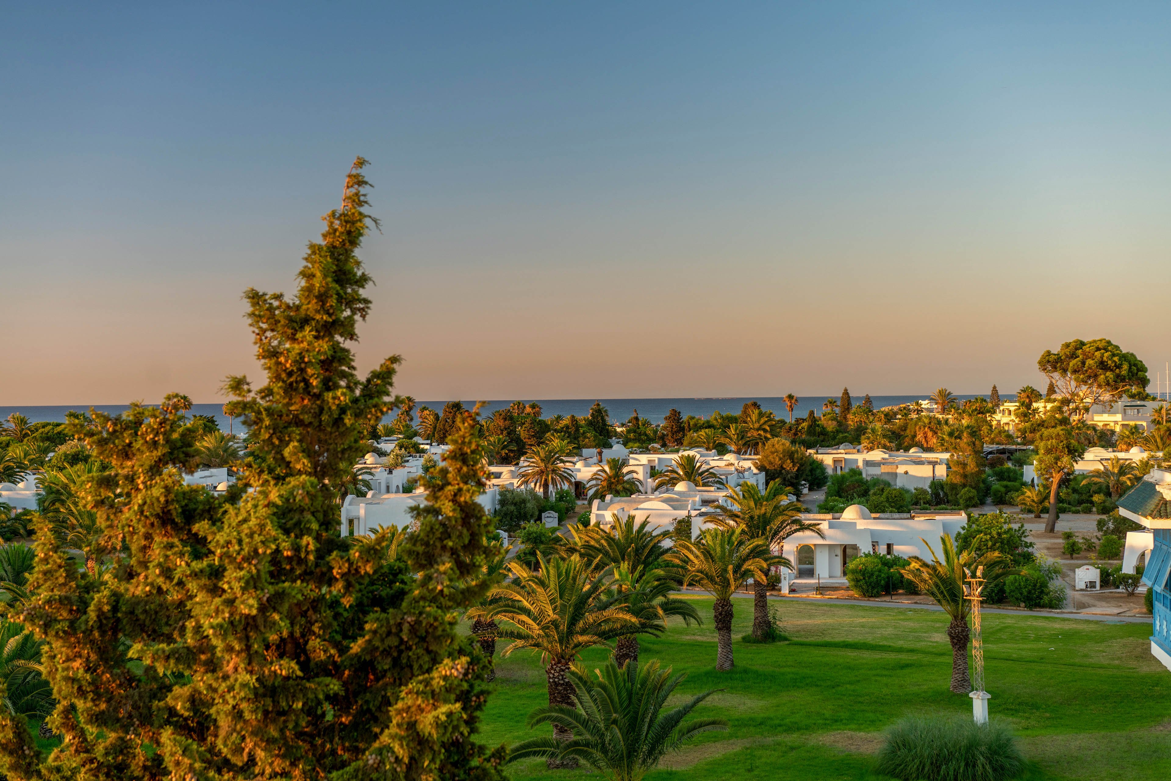 A coastal resort landscape features white low-rise buildings surrounded by lush greenery and palm trees. The scene is captured during golden hour, with warm sunlight casting soft shadows across the lawns. In the distance, the calm sea meets a clear sky that transitions from blue to pastel tones. The overall atmosphere is tranquil and inviting, suggesting a relaxing vacation destination.