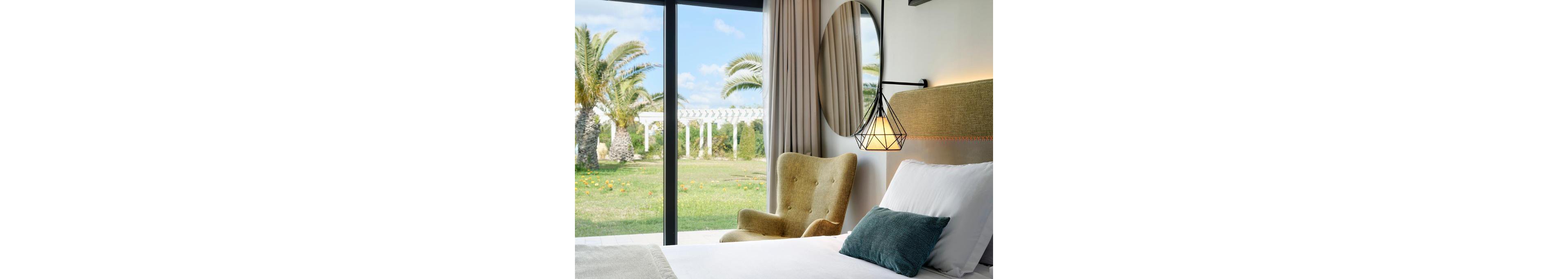 A modern hotel bedroom features a neatly made bed with white linens and a green accent pillow. Beside the bed is a textured armchair and a geometric pendant light hanging from the wall. Large floor-to-ceiling windows open onto a lush garden with palm trees and a white pergola under a blue sky. The scene feels bright, airy, and inviting, suggesting a relaxing vacation setting.