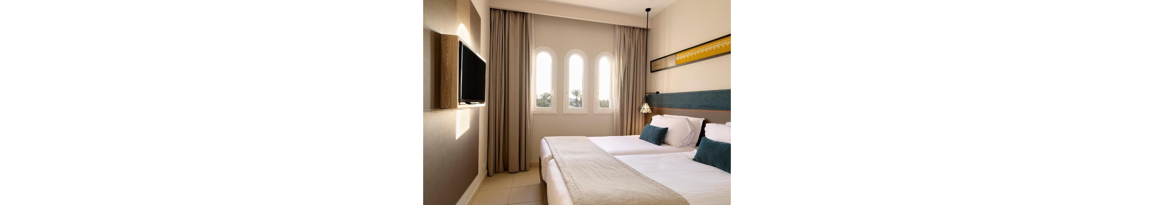 A modern hotel room features two neatly made twin beds with white linens and teal accent pillows. Soft natural light enters through three tall arched windows framed by beige curtains. A flat-screen television is mounted on the wall opposite the beds, and a hanging bedside lamp adds a warm glow. The decor is minimalist with neutral tones and clean lines, creating a calm and inviting atmosphere.