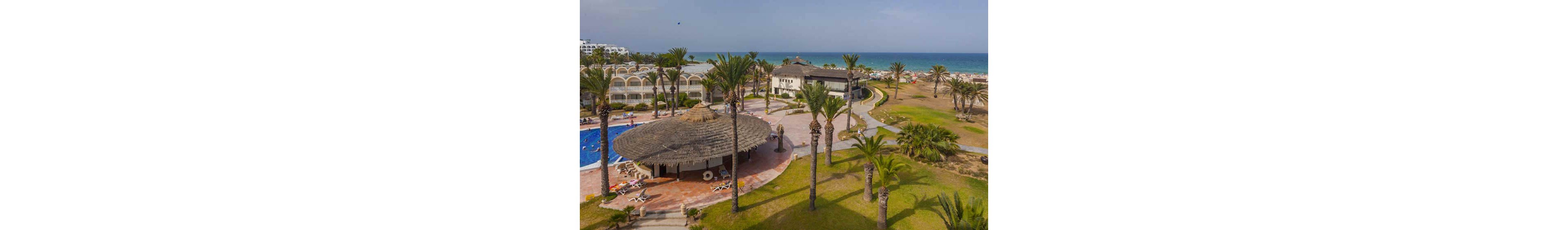 A coastal resort complex is shown from an elevated viewpoint, featuring a large circular thatched-roof bar area beside a blue swimming pool. Palm trees and manicured lawns stretch across the property, leading toward a sandy beach and calm sea in the background. Several low-rise hotel buildings with arched balconies line one side of the scene, while sun loungers are scattered around the pool and near the shoreline. The overall atmosphere is relaxed and summery, suggesting a vacation destination by the ocean.