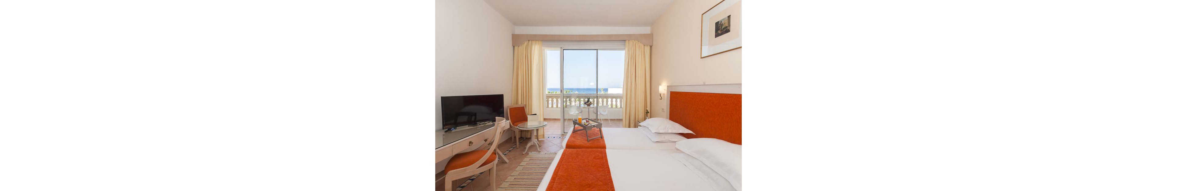 A tidy hotel room features twin beds with orange accents and white linens. A flat-screen television sits on a desk beside a chair and small round glass table. Large sliding doors open to a balcony with balustrade, overlooking a blue sea and clear sky. Soft beige walls, light curtains, and simple decor create a calm, inviting atmosphere.
